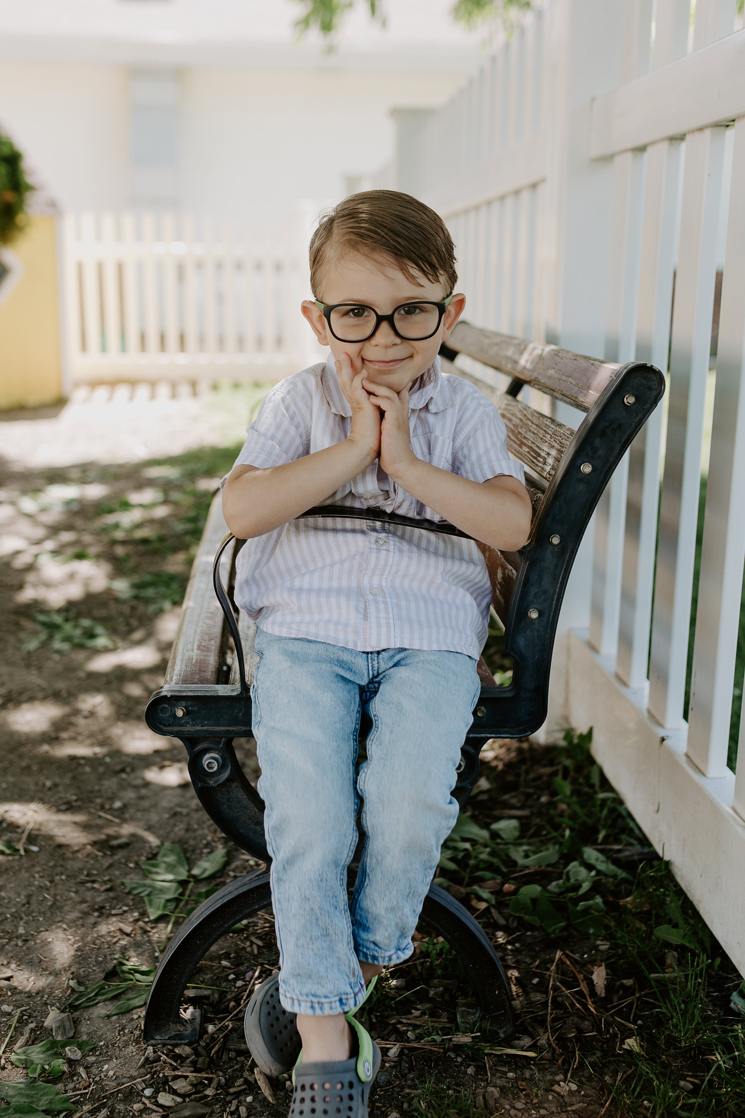 A young boy with glasses, wearing a striped shirt and jeans, sitting on a park bench outdoors, resting his chin on his hands, smiling.