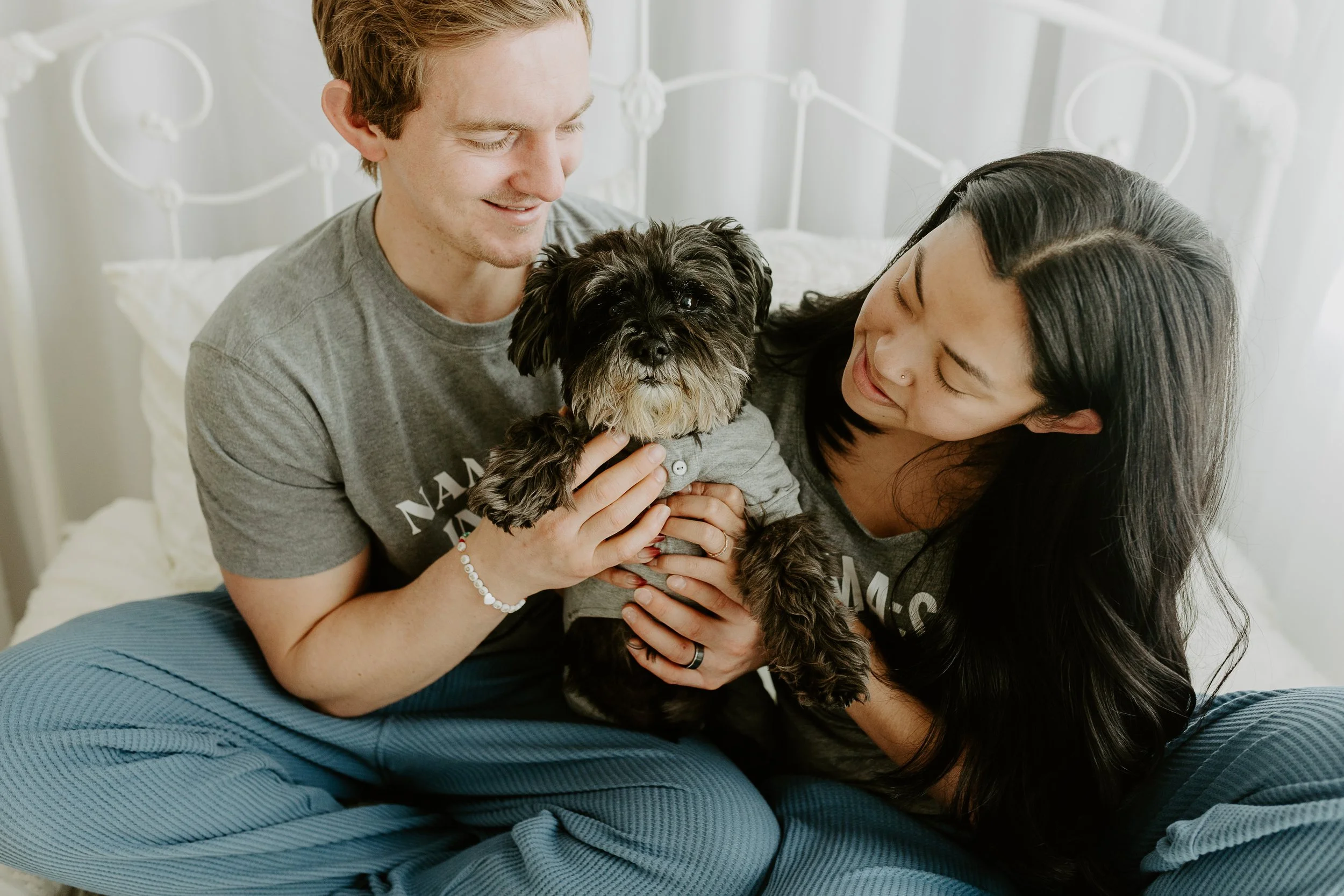 Husband and wife hold their little black dog in-between them while sitting on a white bed
