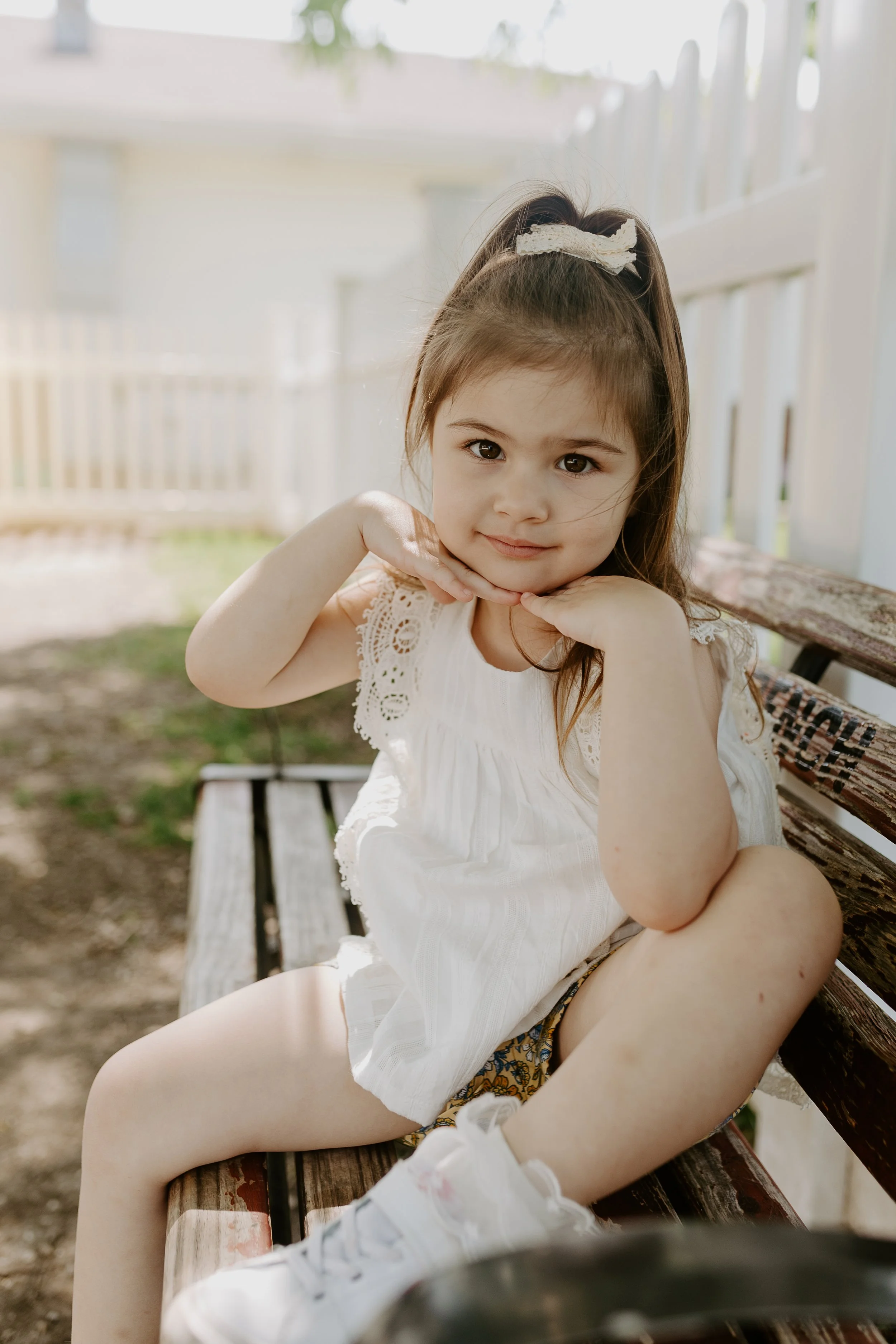 A young girl with a white dress and floral shorts sitting on a wooden bench outdoors, smiling softly with her chin resting on her hands, wearing a hair bow.
