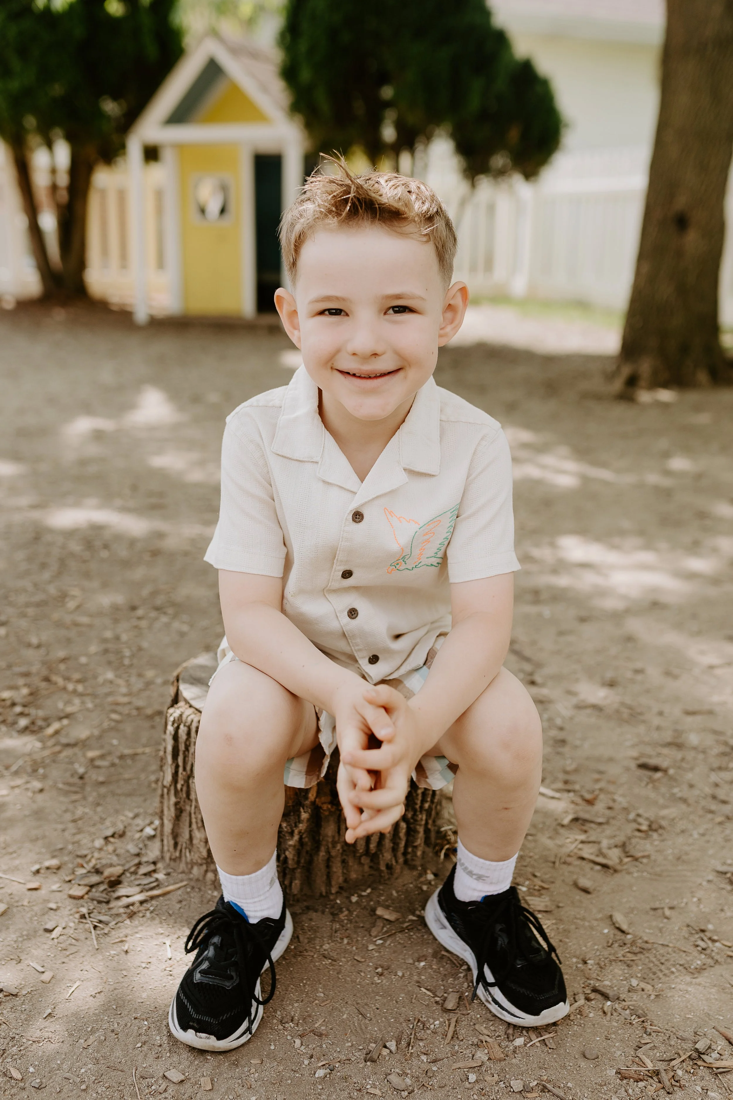 A young boy sitting on a tree stump in a backyard, smiling at the camera. He is wearing a beige short-sleeve shirt with a colorful embroidered bird, shorts, white socks, and black sneakers. There are trees, a small yellow playhouse, and a white picket fence in the background.