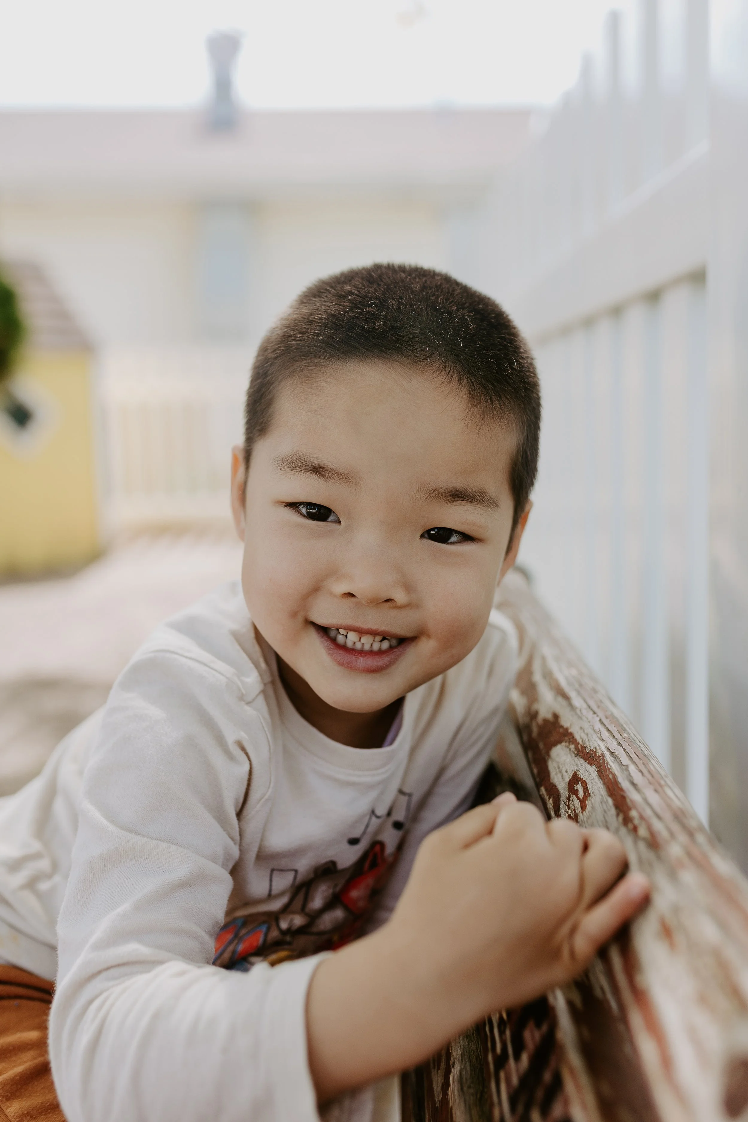 A young boy with short hair smiling and looking at the camera, leaning on a wooden surface outdoors.