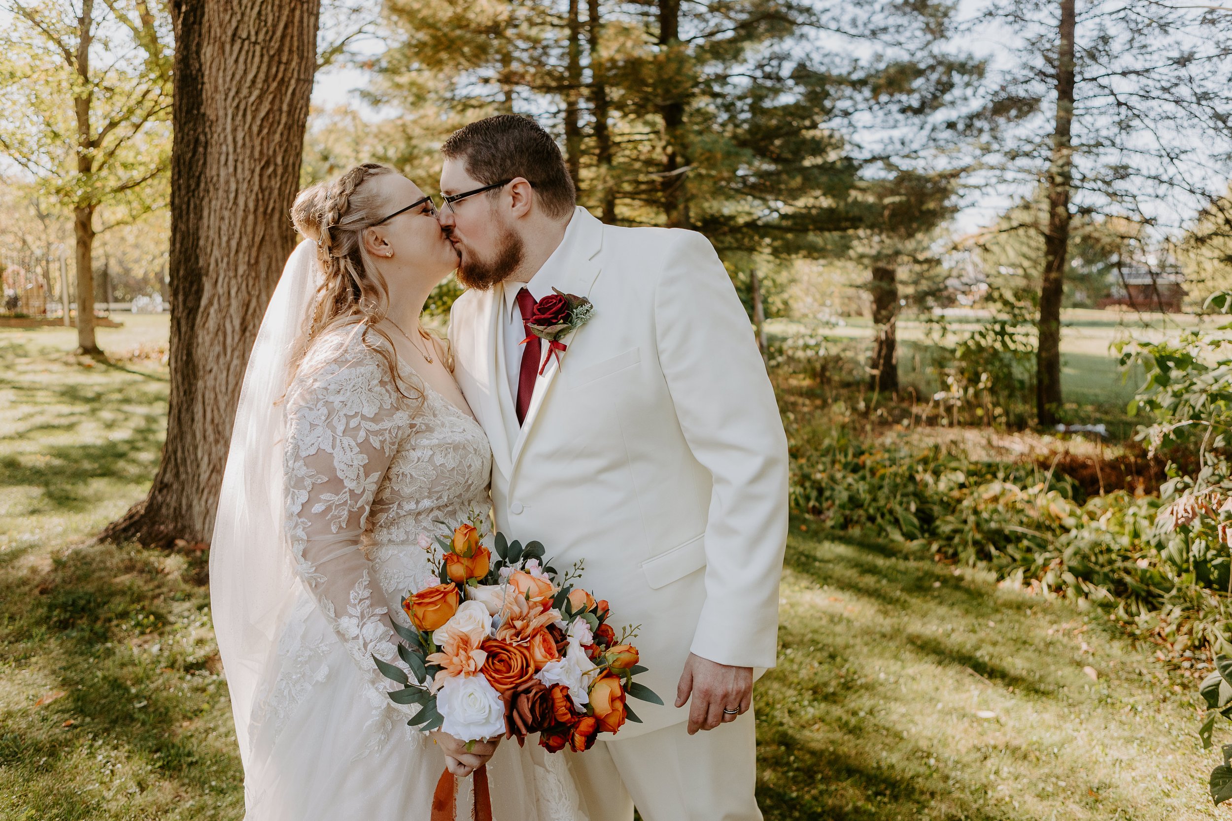 A newly married couple shares a kiss outdoors during daytime, the bride wearing a lace wedding dress and holding a bouquet of orange and white flowers, and the groom in a white suit with a red tie and boutonniere, surrounded by trees in a park-like setting.