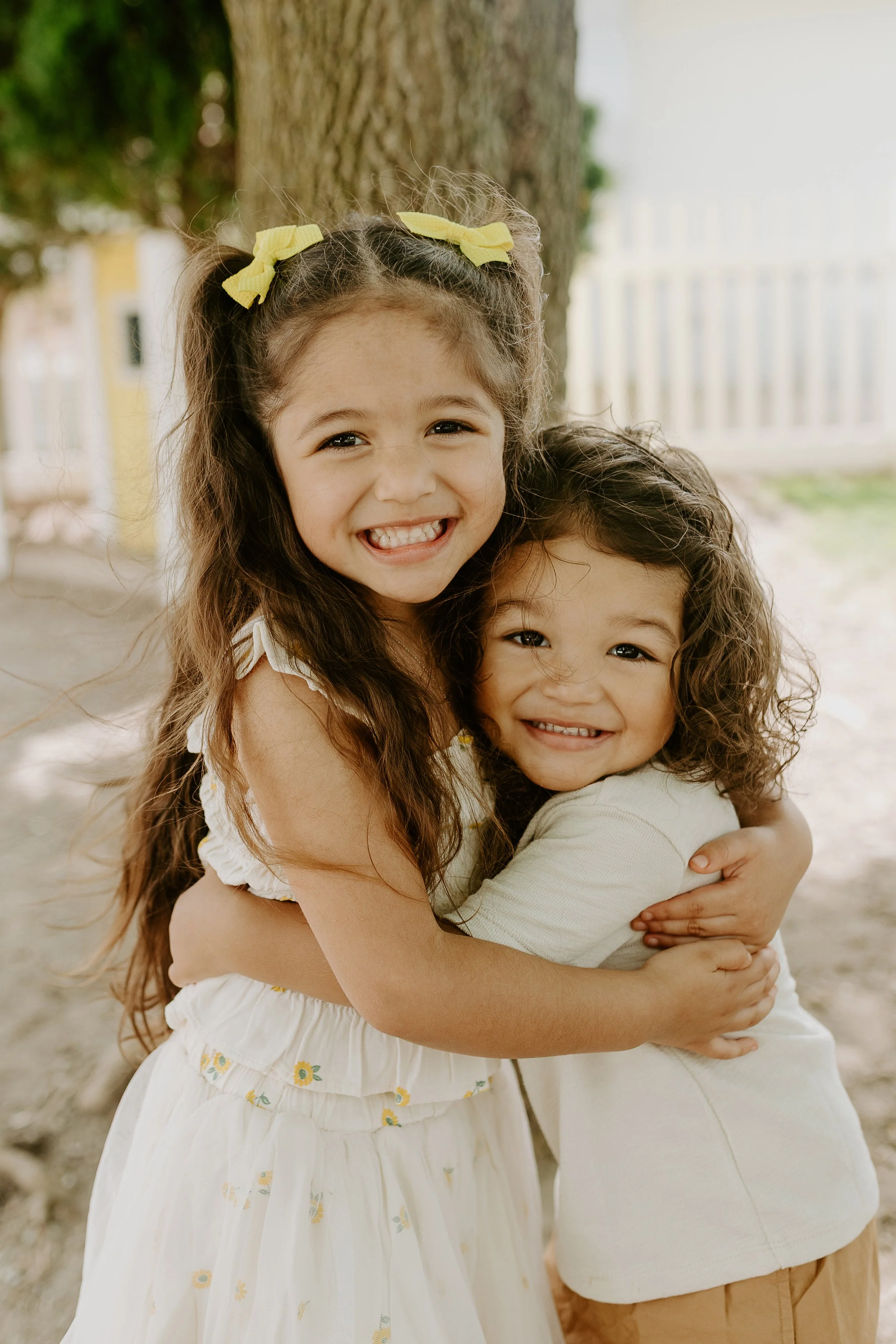 Two young girls hugging outdoors near a tree, smiling happily.