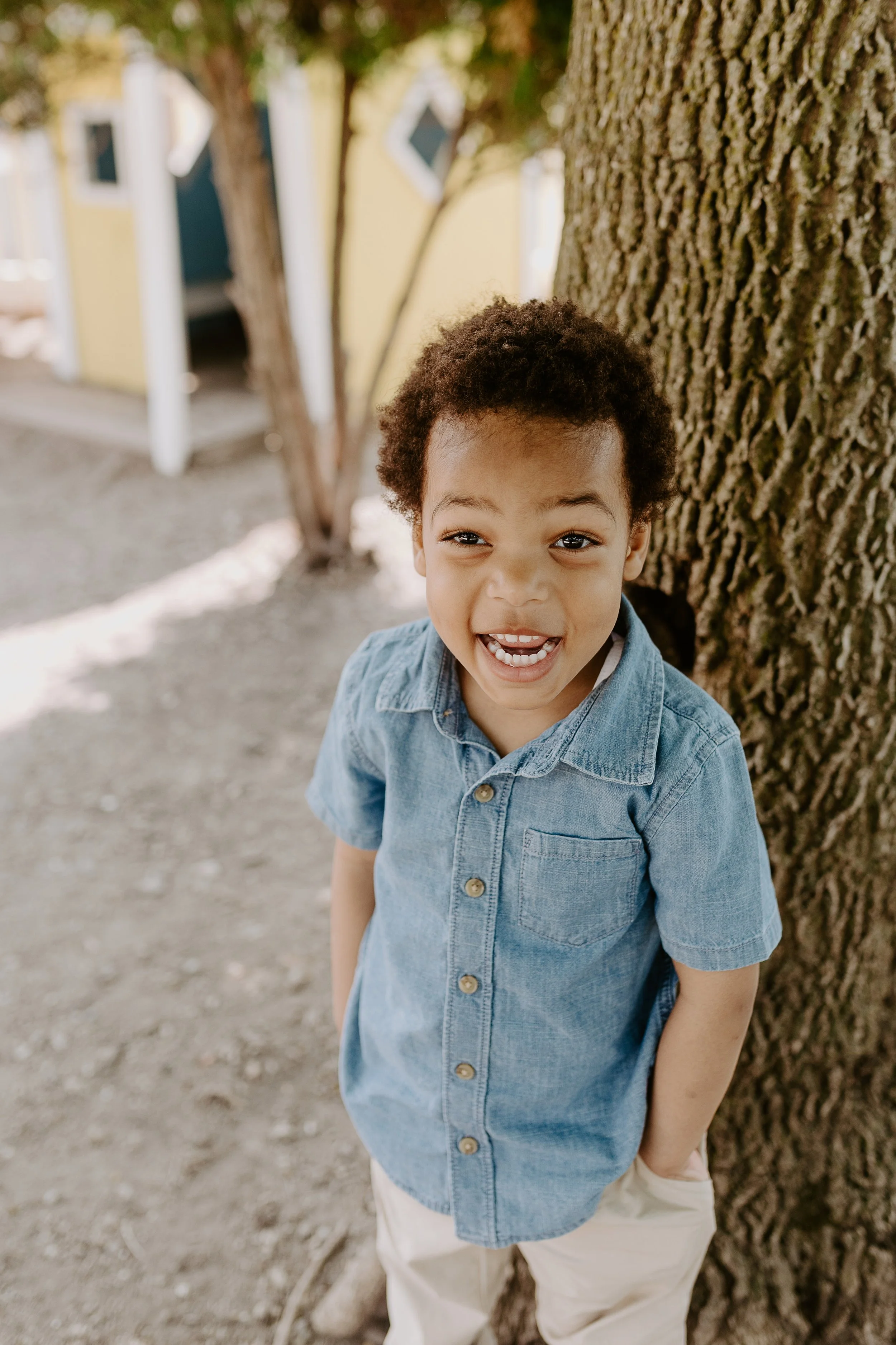 A young boy with curly dark hair and a big smile, wearing a denim shirt and light-colored pants, standing outdoors leaning against a large tree.