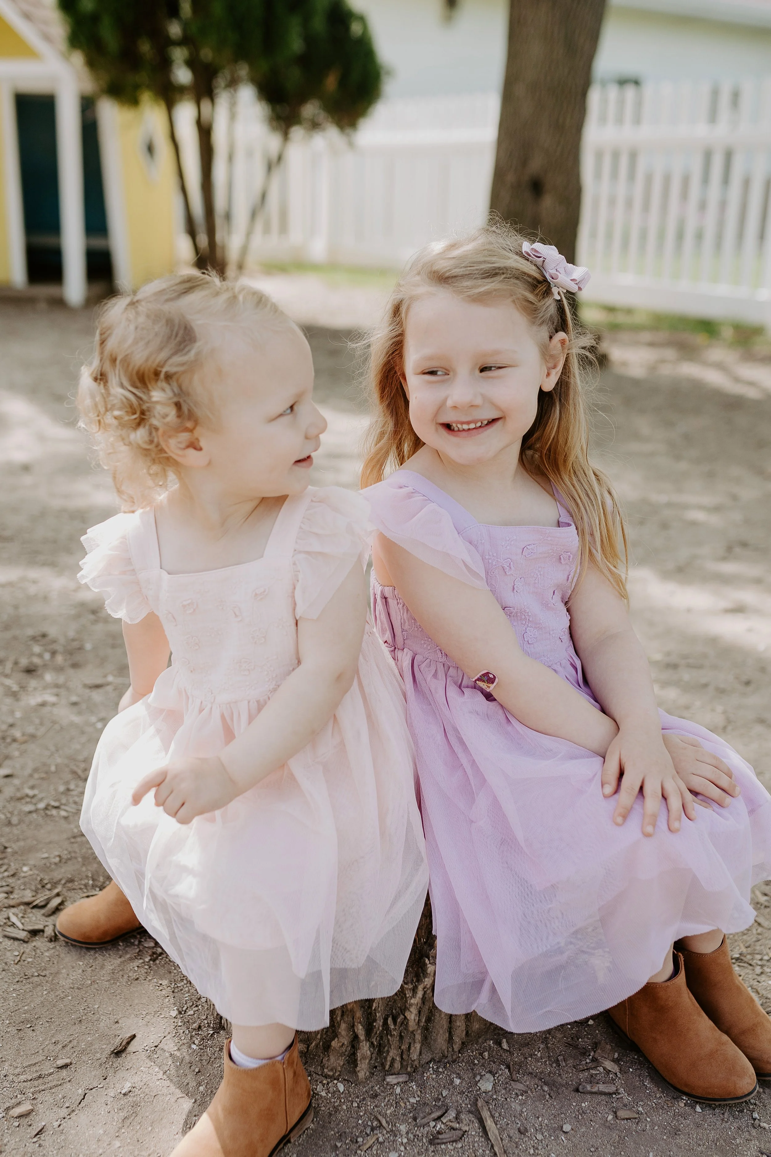 Two young girls sitting outdoors on a tree stump, dressed in pastel-colored dresses, smiling and talking to each other.