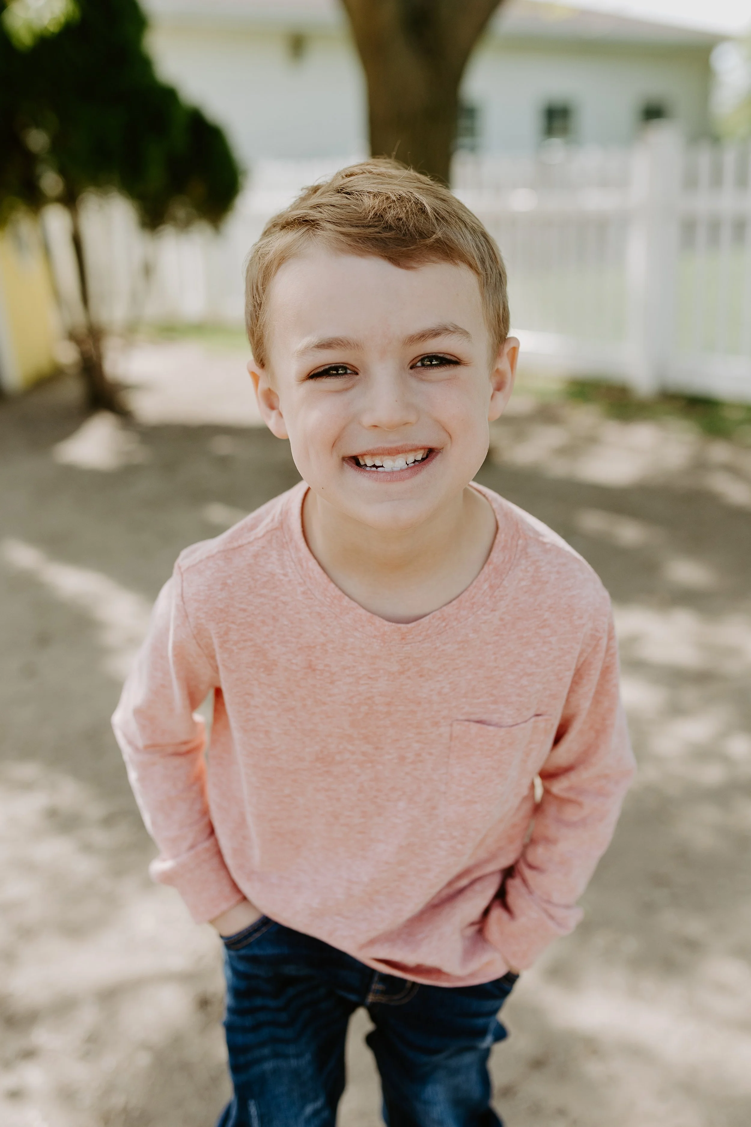 A smiling boy with light brown hair, wearing a peach long-sleeve shirt and dark blue jeans, standing outdoors with trees, a white fence, and a house in the background.