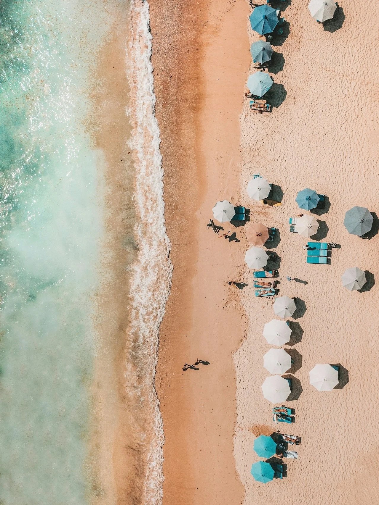 Aerial view of a beach with turquoise water, sandy shore, and colorful umbrellas with people walking and relaxing on the beach.