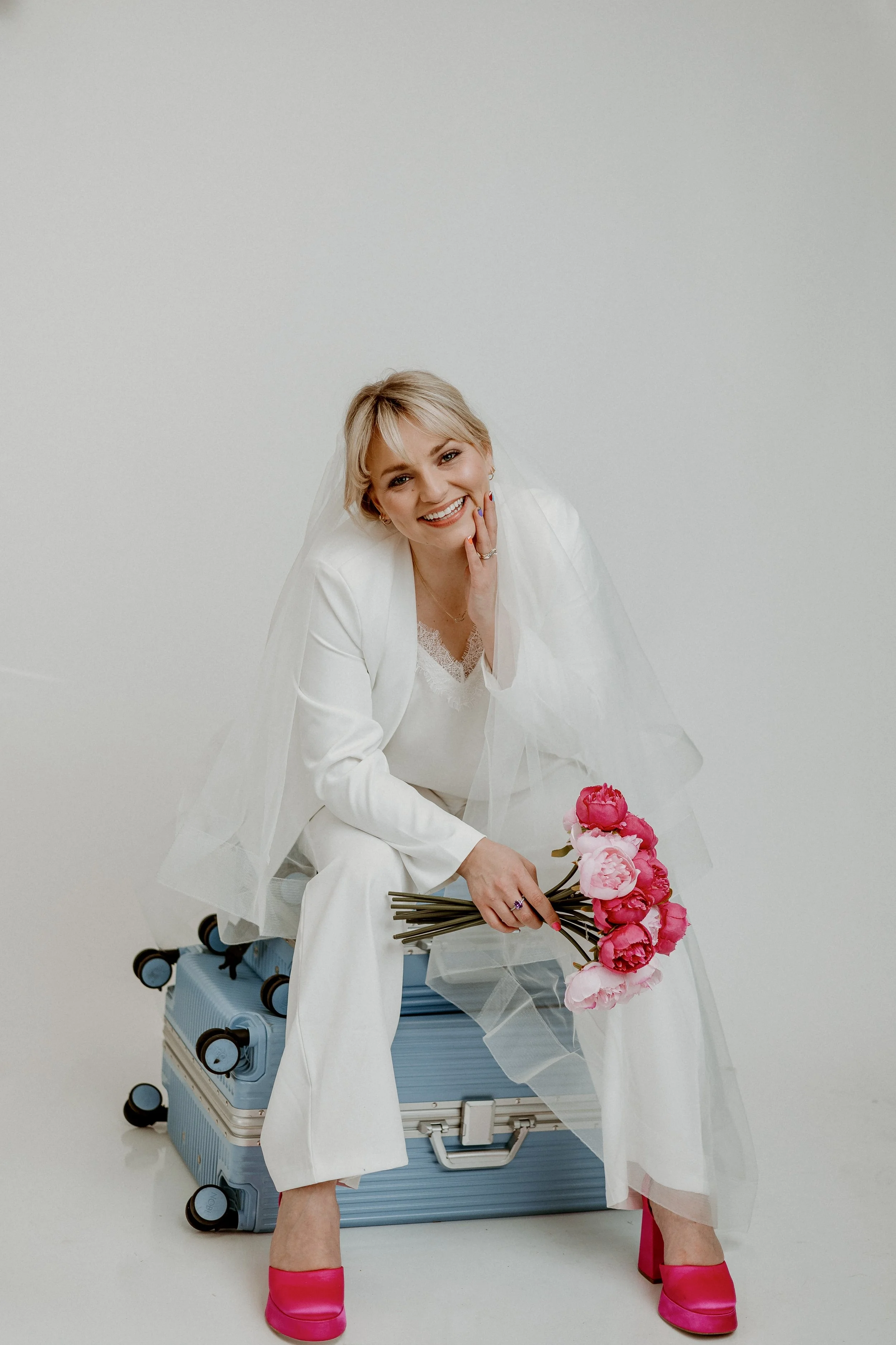 A woman dressed in white sitting on a blue suitcase, holding pink peony flowers, smiling, with pink shoes, against a plain white background.