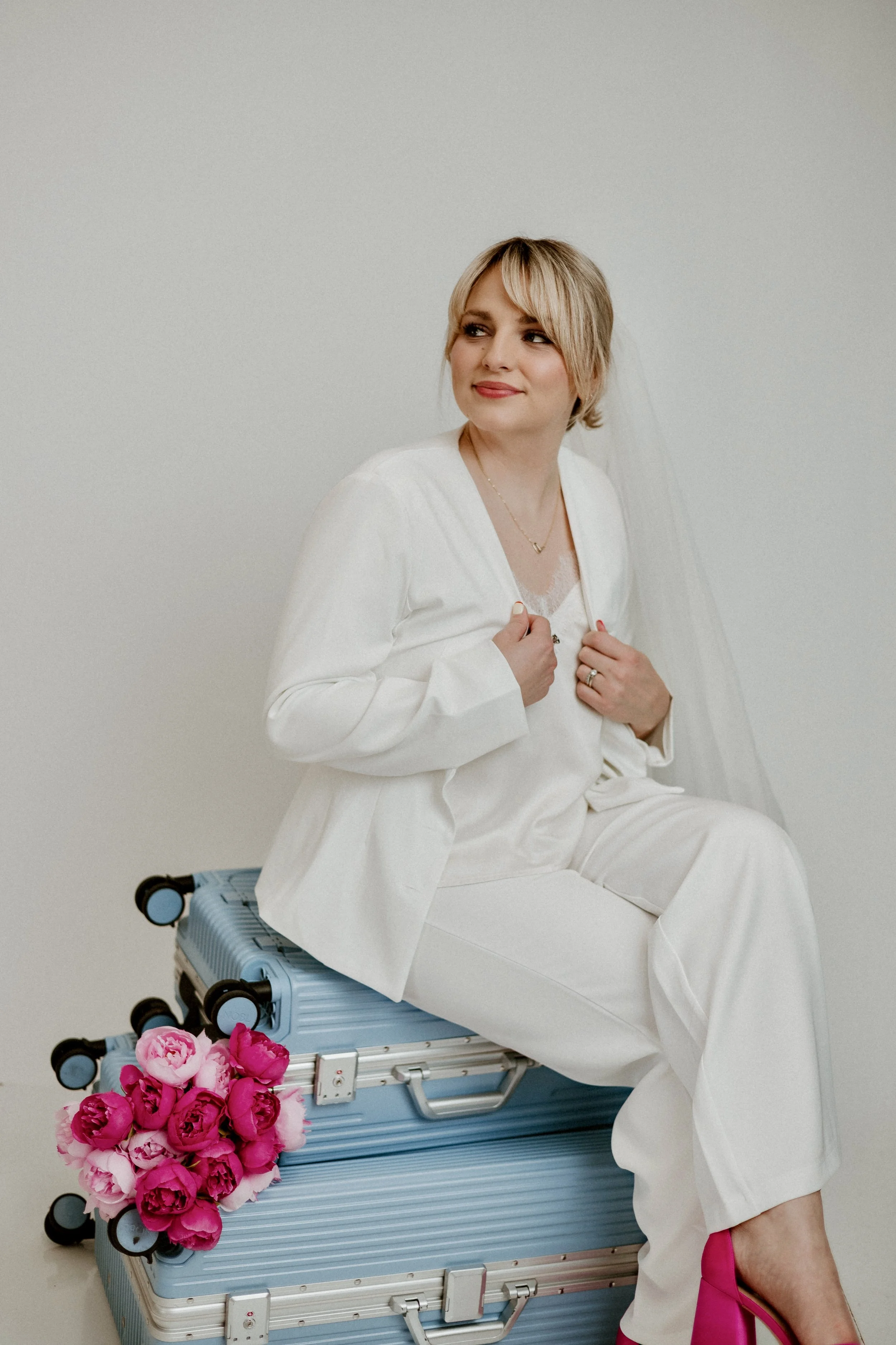 A woman dressed in white sitting on a blue suitcase with pink flowers and pink high heels, against a plain white background.
