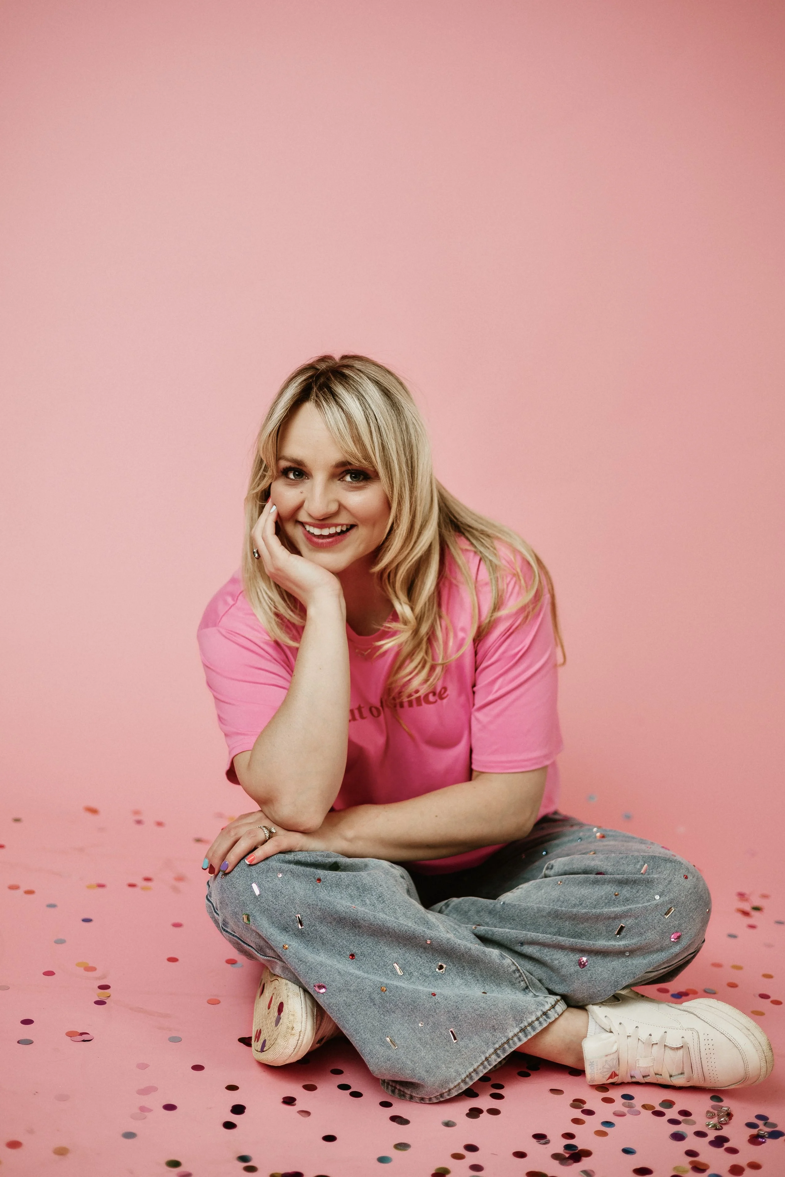 A woman with blonde hair sitting on a pink background, smiling, wearing a pink shirt and gray pants with confetti, surrounded by scattered confetti on the floor.