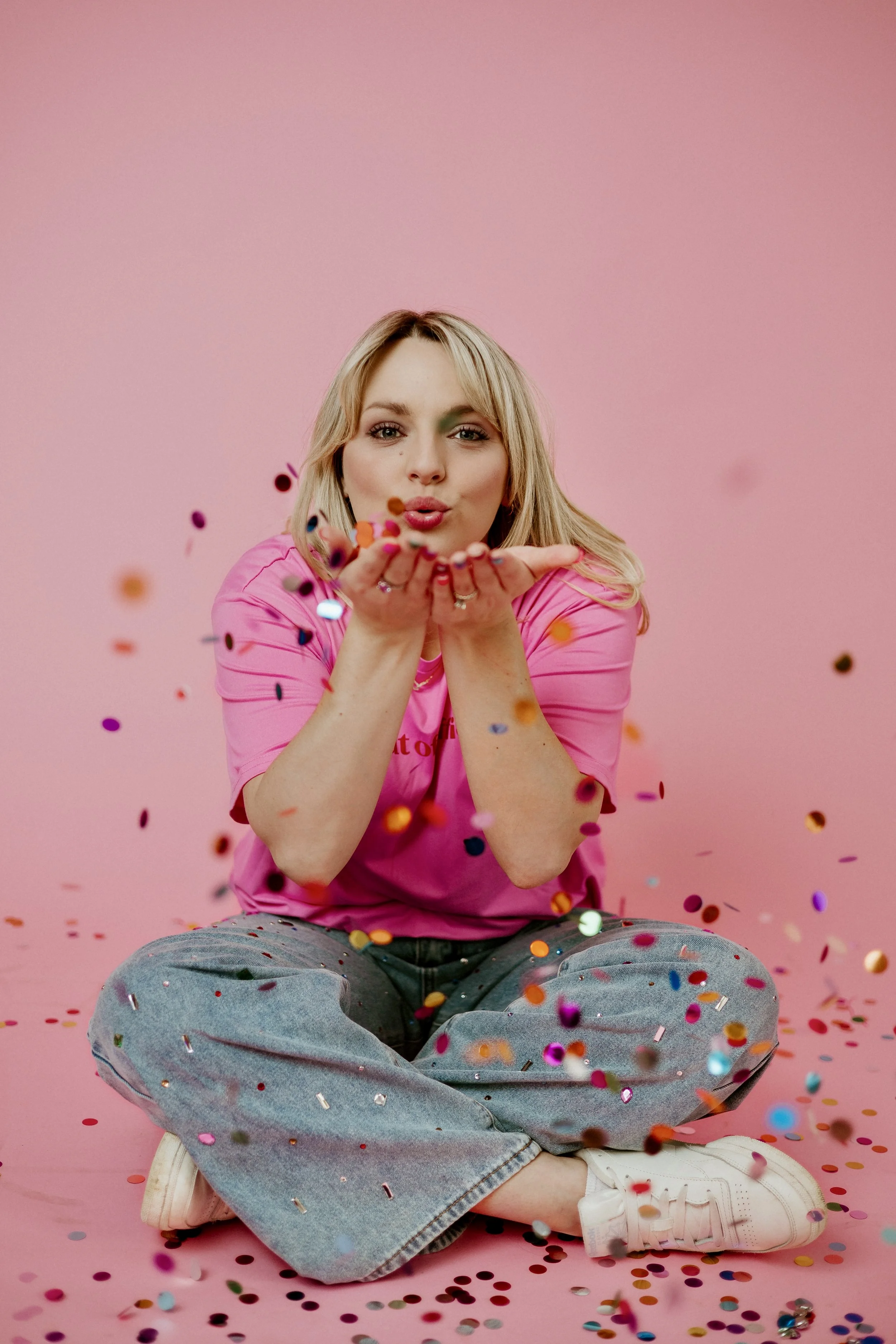 A young woman sitting cross-legged in front of a pink background, blowing colorful confetti towards the camera.