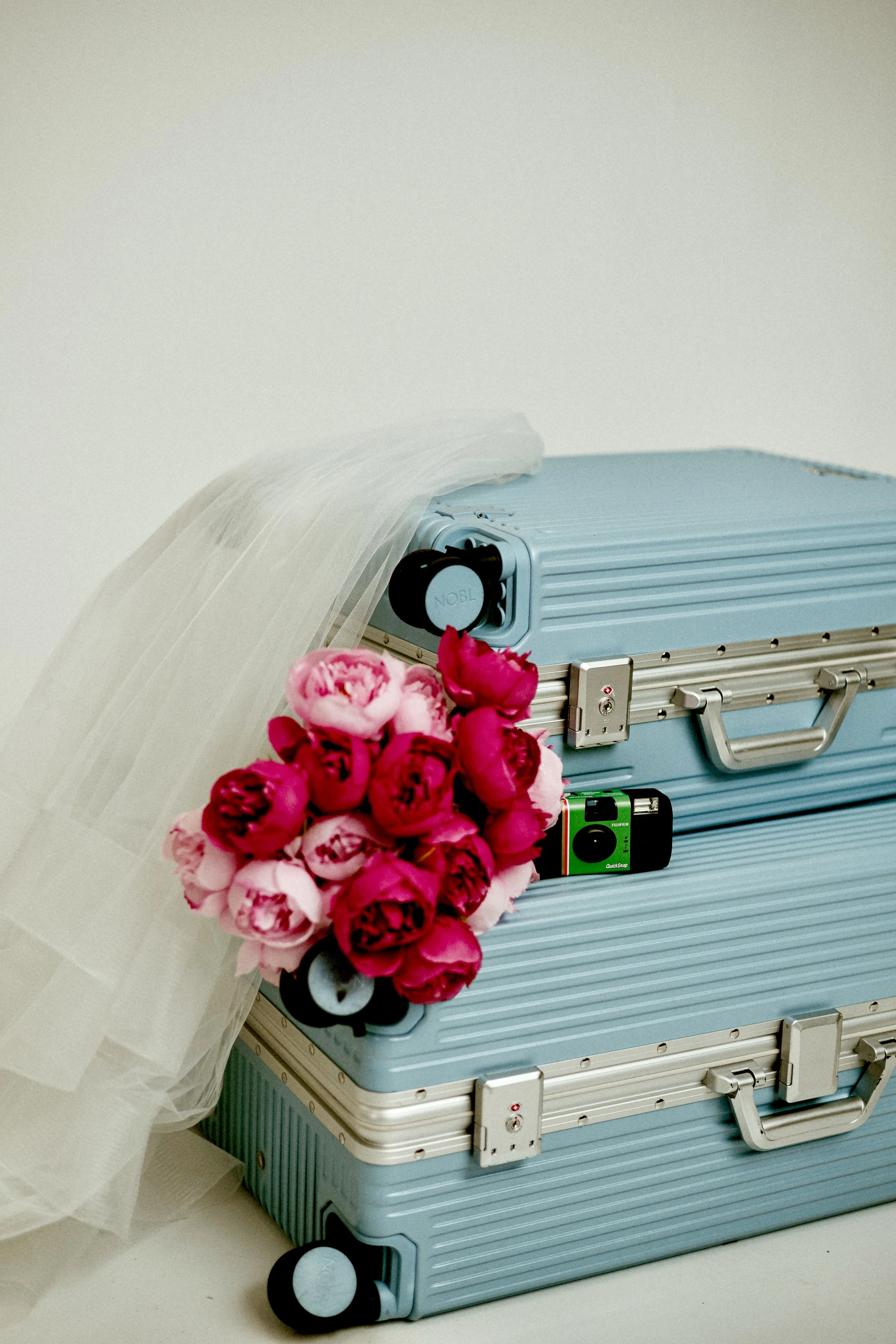 A stack of two blue suitcases with a bunch of pink and red peony flowers and a piece of white tulle fabric draped over the top suitcase.