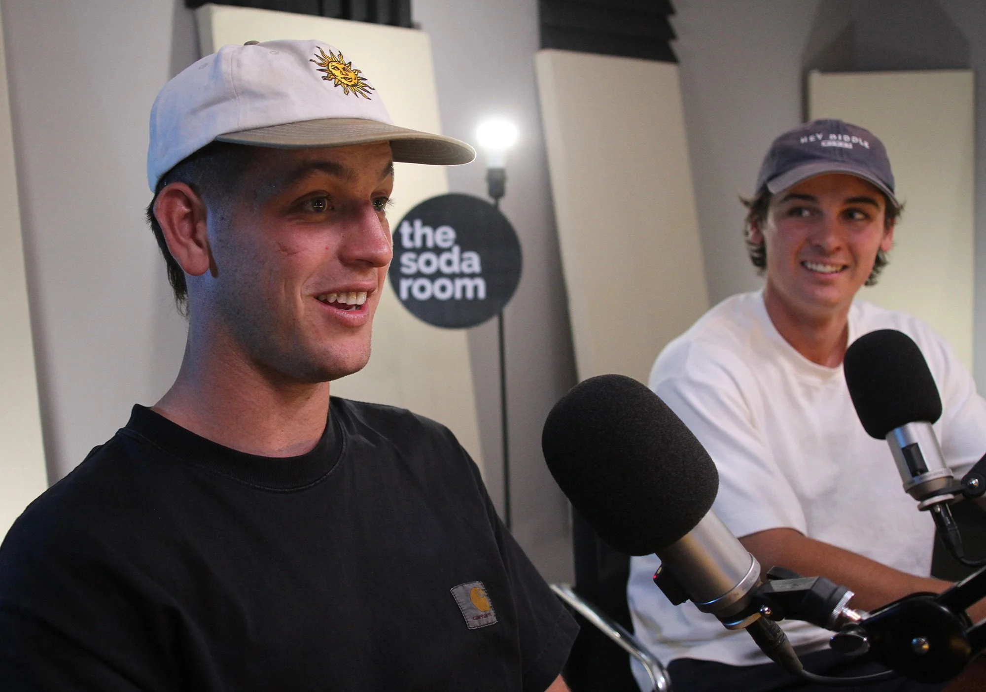 Two young men sit at microphones during a radio interview at 'the soda room'. The man on the left wears a beige cap with a sun and moon design and a black t-shirt, while the man on the right wears a gray cap and a white t-shirt, both smiling.