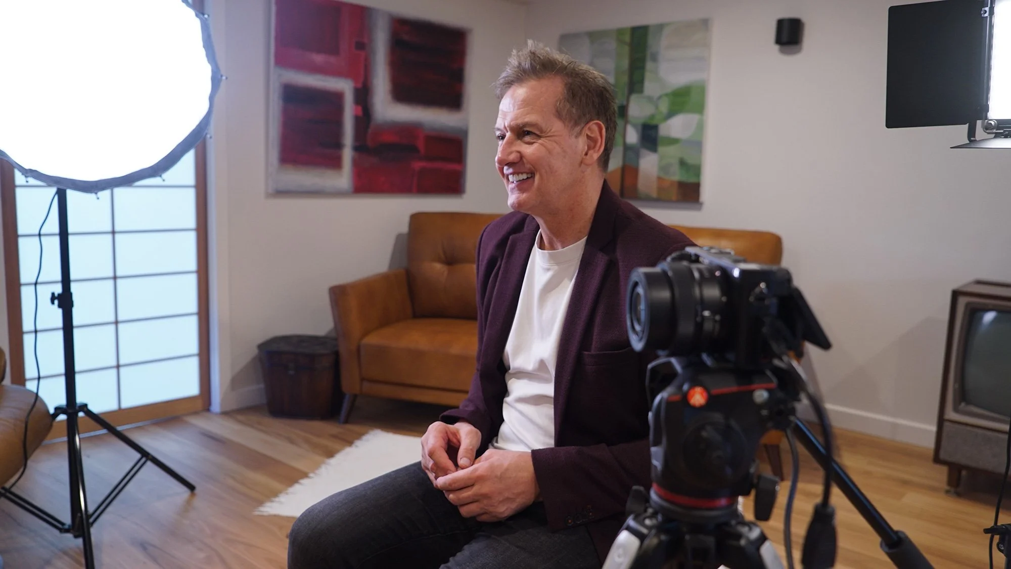 A man sitting in a recording studio, smiling, with lighting and camera equipment set up for filming.