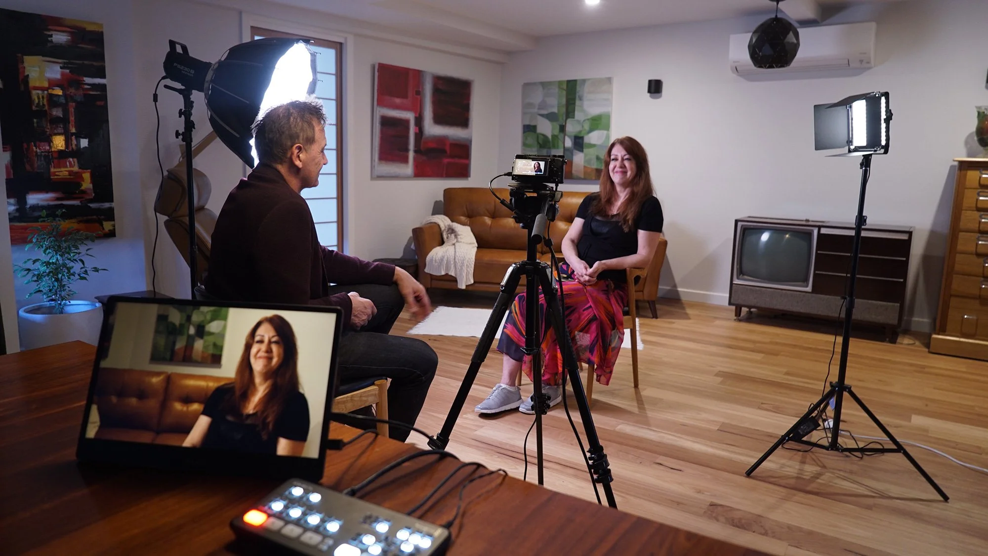 A woman is sitting on a wooden chair in a living room, during a video recording. She is smiling and wearing a black top and colorful skirt. Two studio lights and a camera on a tripod are set up around her. A man is sitting on another chair facing her, talking. A monitor on a table shows her image, and a small lighting control device is nearby. The room has artwork on the walls, a vintage TV stand with an old TV, and wooden flooring.