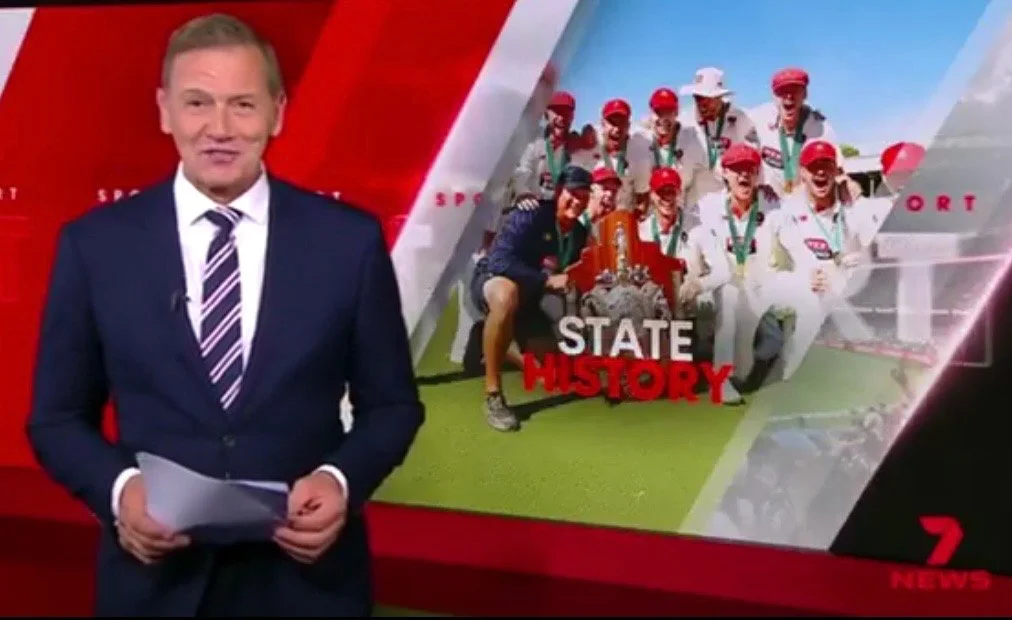 News anchor standing in front of a graphic showing a sports team celebrating with medals and a trophy, with the words "State History" and "7 News" in the background.