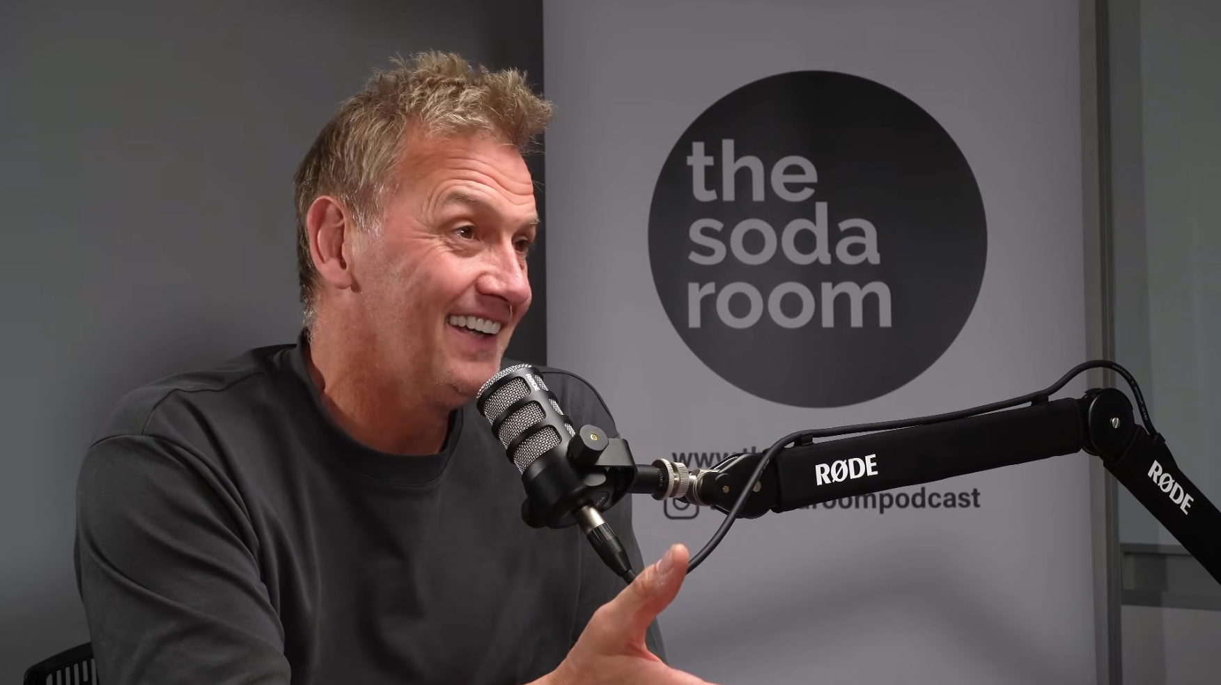 A man with curly hair and a black shirt speaking into a professional microphone in a recording studio with a sign reading 'the soda room' in the background.