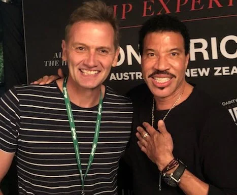 Two men smiling and posing for a photo together at an event, one wearing a striped shirt and the other wearing a black shirt with a necklace.