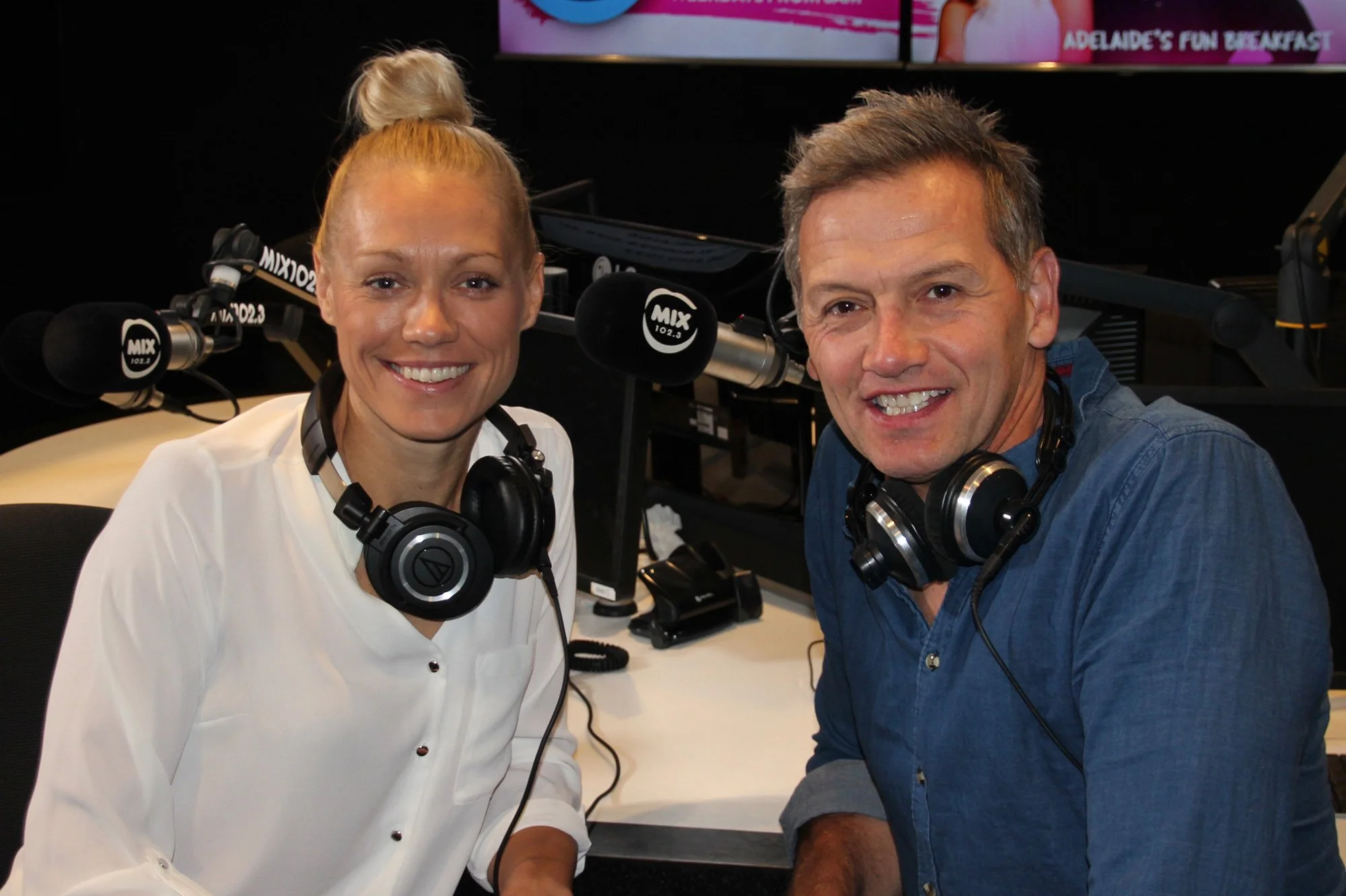 Two radio hosts, a woman in a white blouse and a man in a blue shirt, sitting in a recording studio with microphones, headphones, and broadcasting equipment.