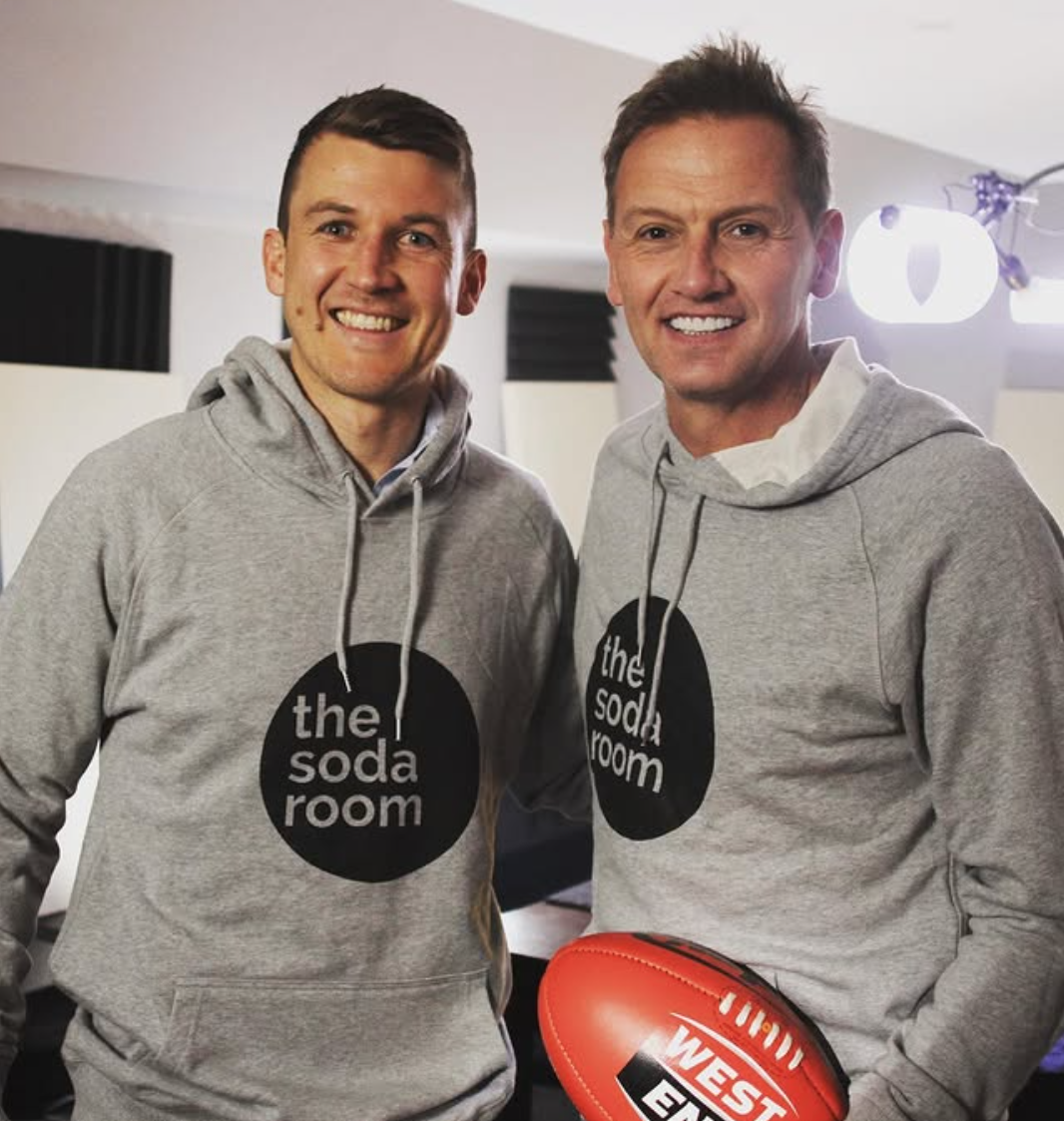Two men smiling, wearing matching gray hoodies with 'the soda room' logo, holding a football in a studio or recording space.