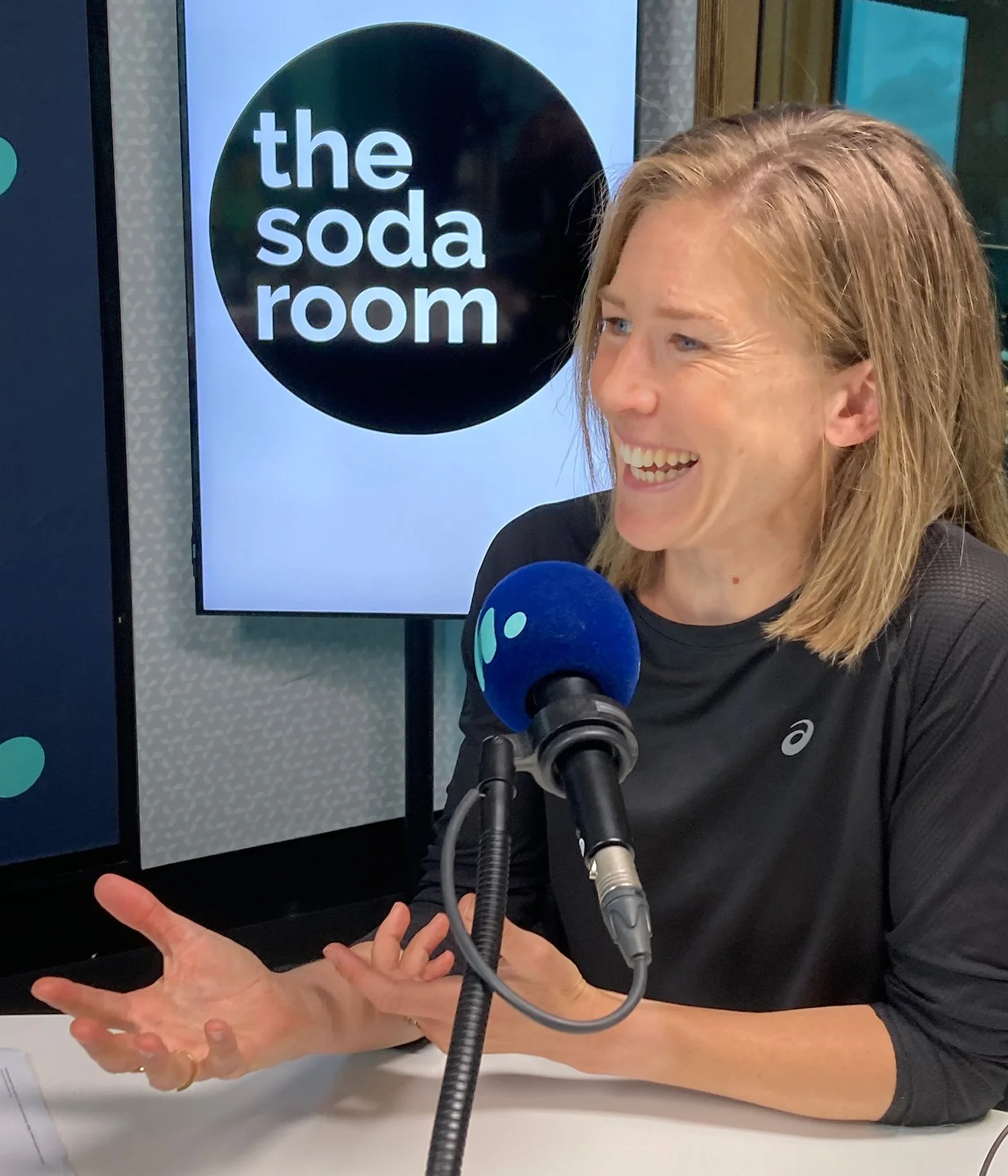 A woman speaking into a microphone during an interview, sitting at a table with a sign behind her that reads 'the soda room'.