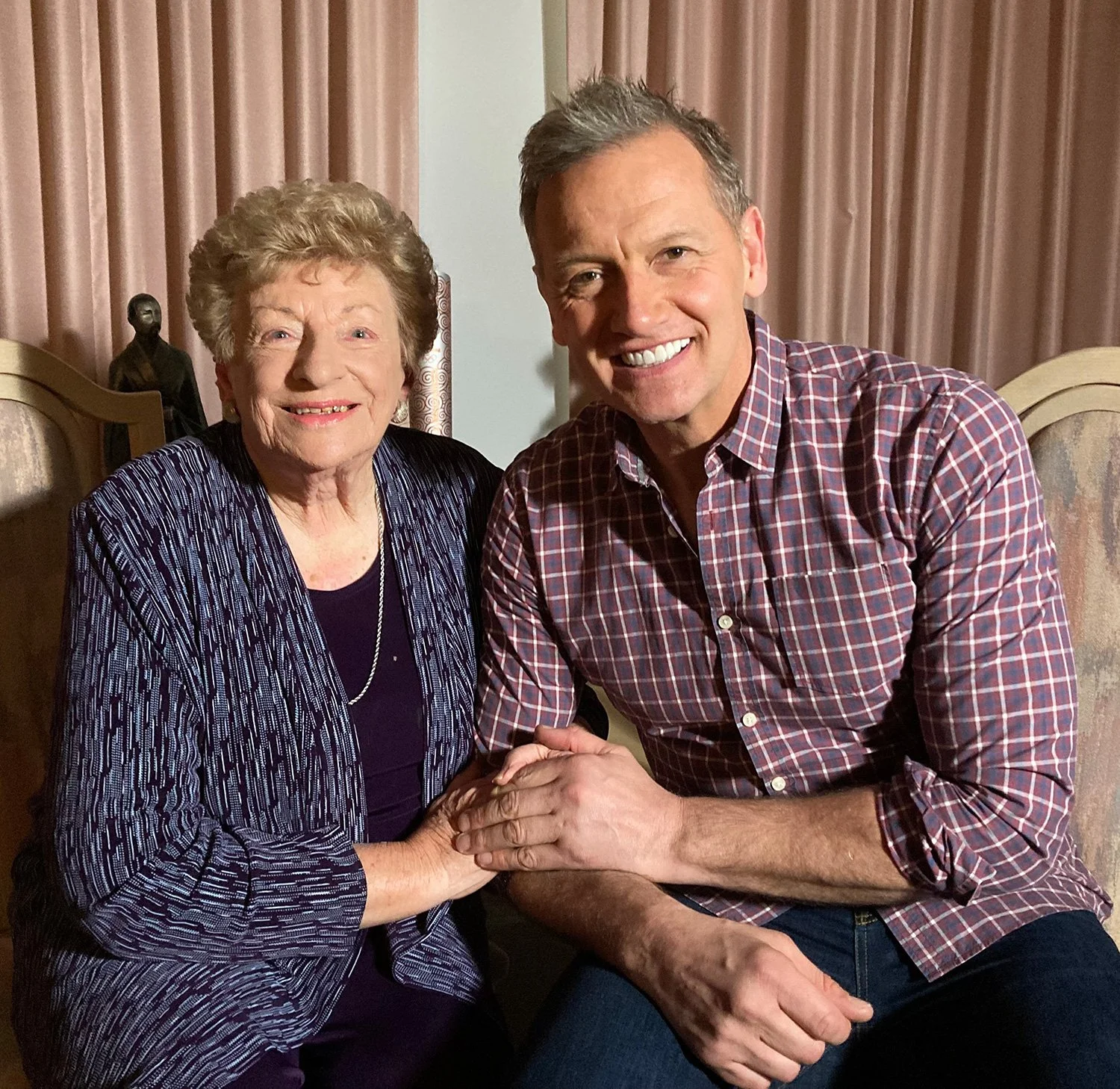 An elderly woman and a middle-aged man sitting together, holding hands, and smiling at the camera in a warmly lit room with pink curtains in the background.