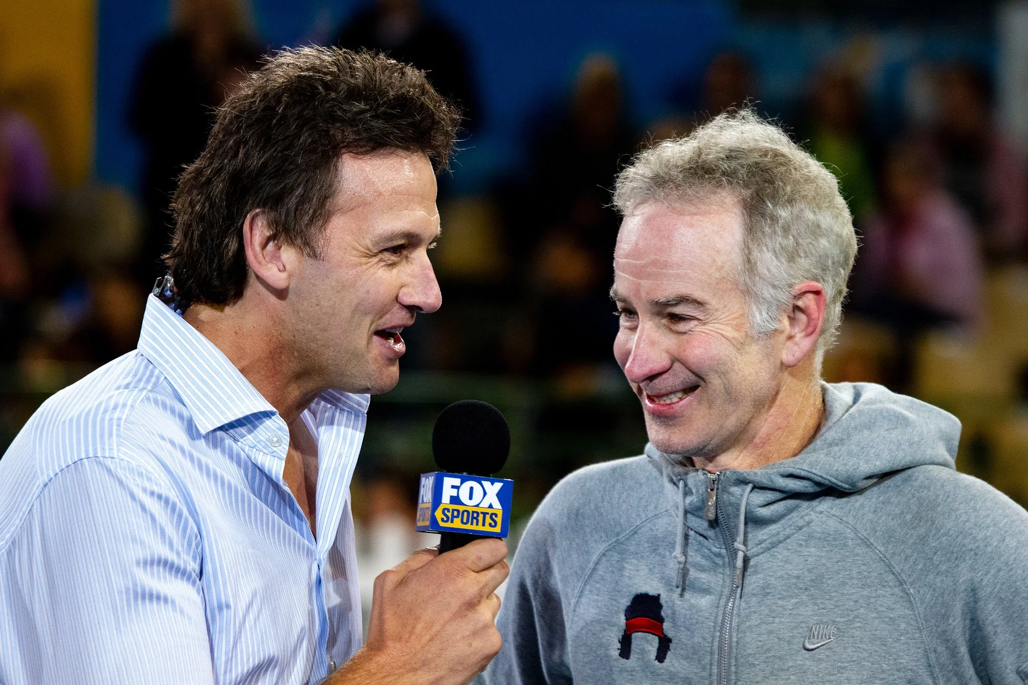 Two men are having a conversation on a sports interview, one is holding a FOX Sports microphone. The man on the left has dark hair and is wearing a light blue striped shirt, while the man on the right has gray hair and is wearing a gray Nike hoodie. They are smiling and facing each other, with a blurred crowd in the background.