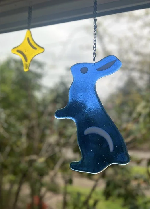 A blue glass penguin hanging from a chain with a yellow star-shaped decoration nearby, outdoors with trees in the background.