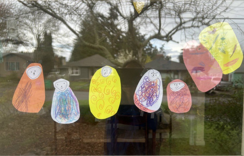 Colorful drawings of abstract characters with smiling faces, made by children, taped to a window with a view of trees and houses outside.