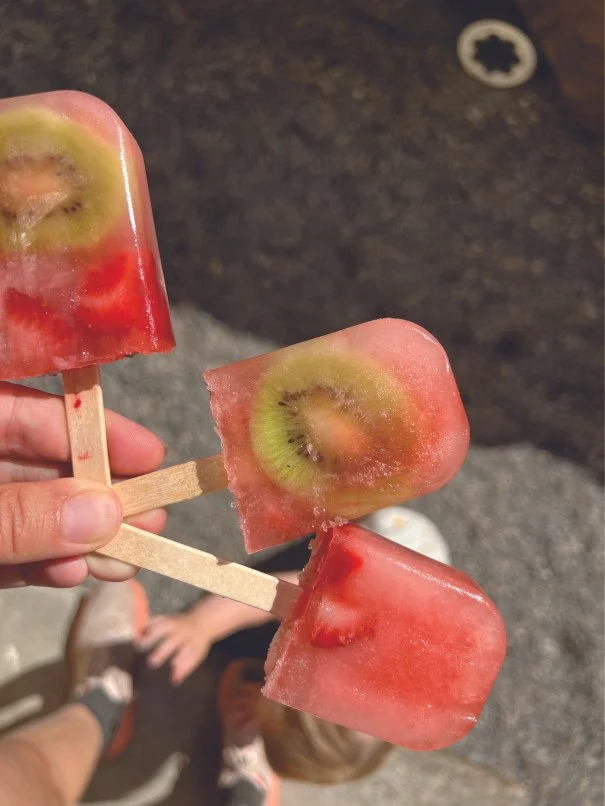 Three watermelon and kiwi fruit popsicles held by a person's hand. The popsicles are melting slightly and are on wooden sticks. The background shows part of a dark surface and a child's legs and feet.