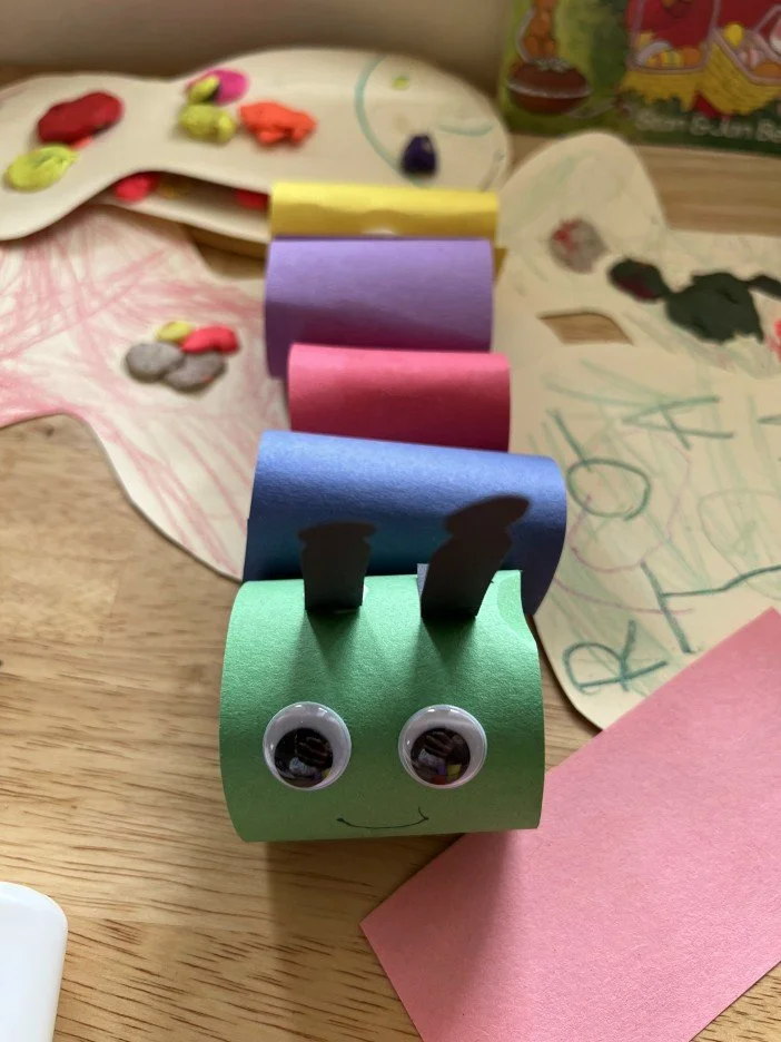 Colorful paper craft of a caterpillar with googly eyes and a smile, on a wooden table surrounded by drawing papers and art supplies.