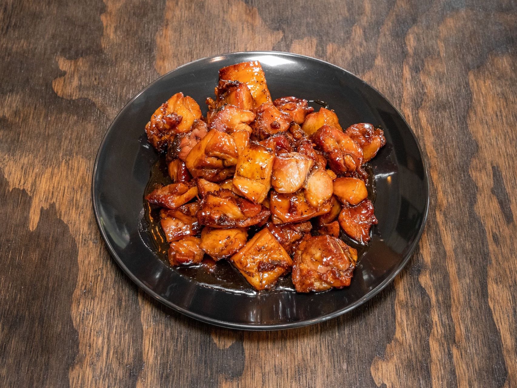 A black plate filled with chunks of glazed and cooked meat on a dark wooden table.