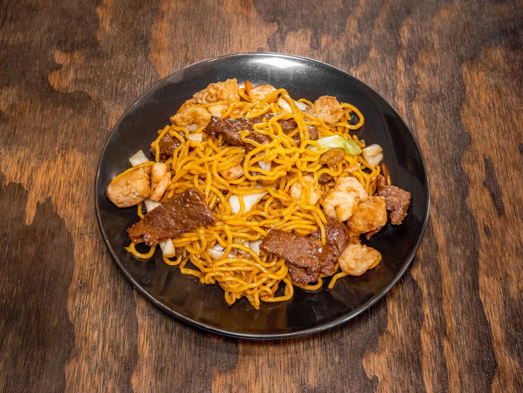 A black plate of stir-fried noodles with shrimp, beef, chicken, and vegetables on a wooden table.