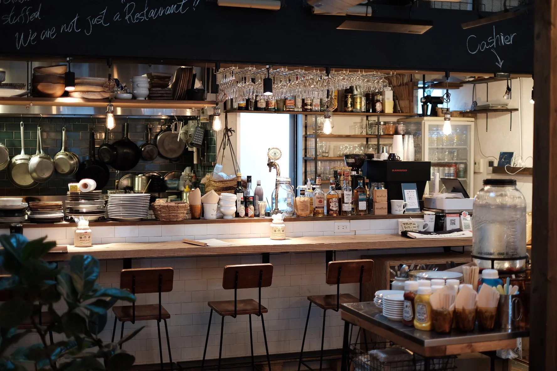 カウンター席の小さなカフェの内装で、キッチンとレジ、コーヒーマシン、瓶入りの酒類、グラス、皿、調理道具などが見える。