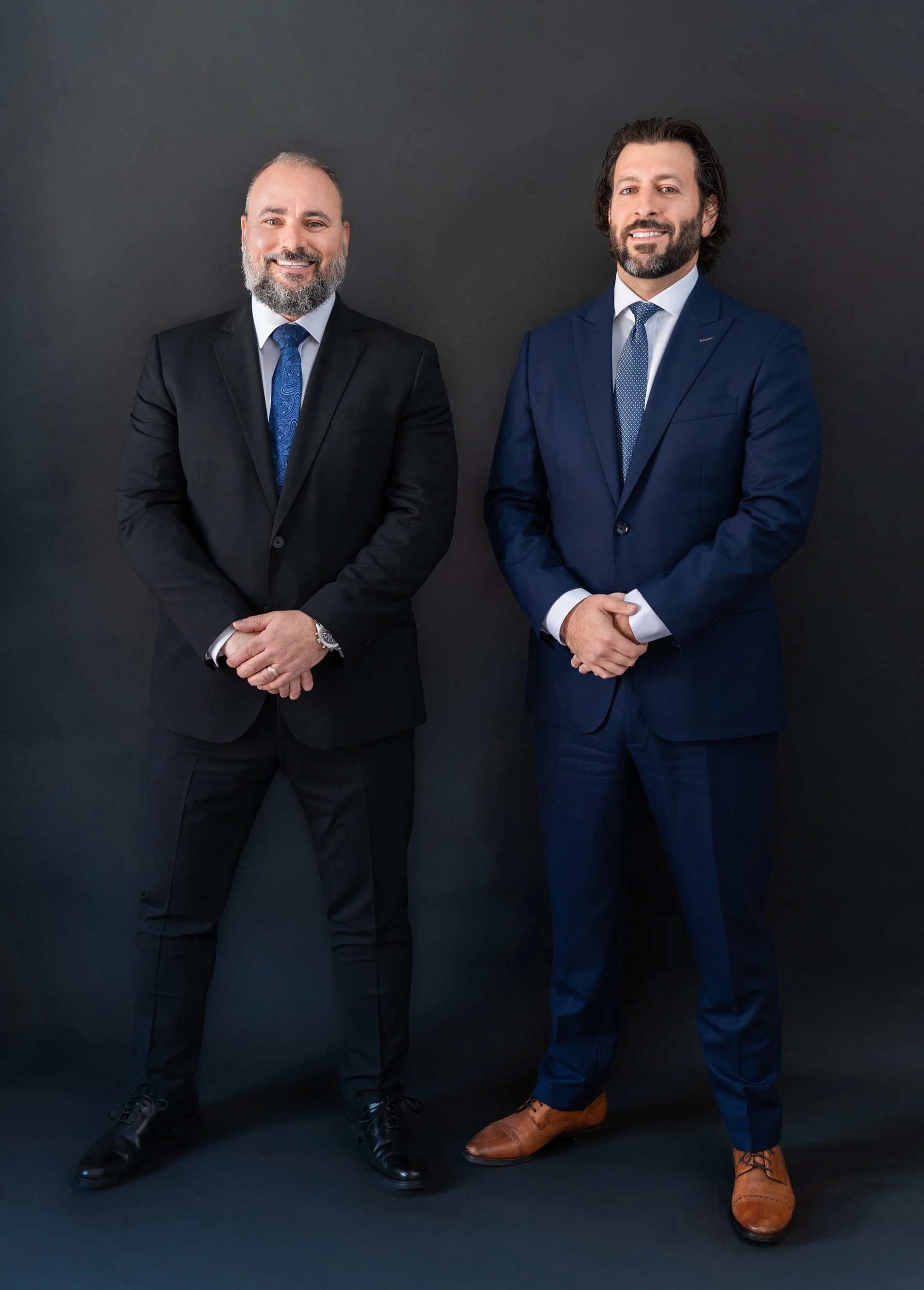 Daniel P. Maltese and Jon-Paul Gabriele dressed in suits are standing against a dark background, smiling, and looking at the camera.