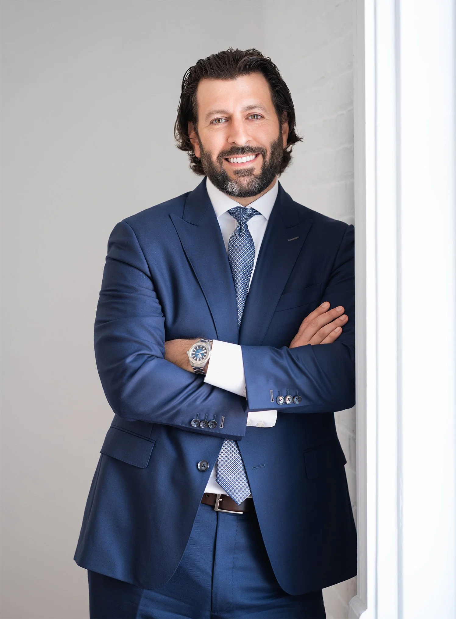 Jon-Paul Gabriele in a dark blue suit, white shirt, and patterned tie stands with crossed arms in front of a plain light-colored wall and a window.
