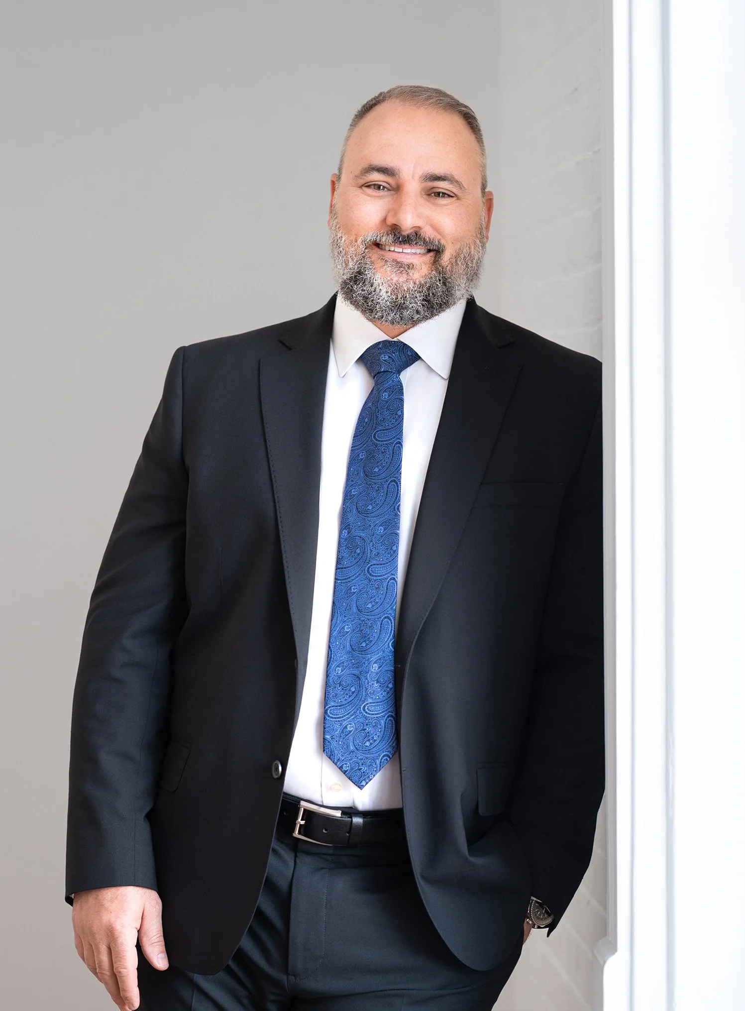 Daniel P. Maltese wearing a black suit, white shirt, and blue tie, smiling and standing indoors near a window.