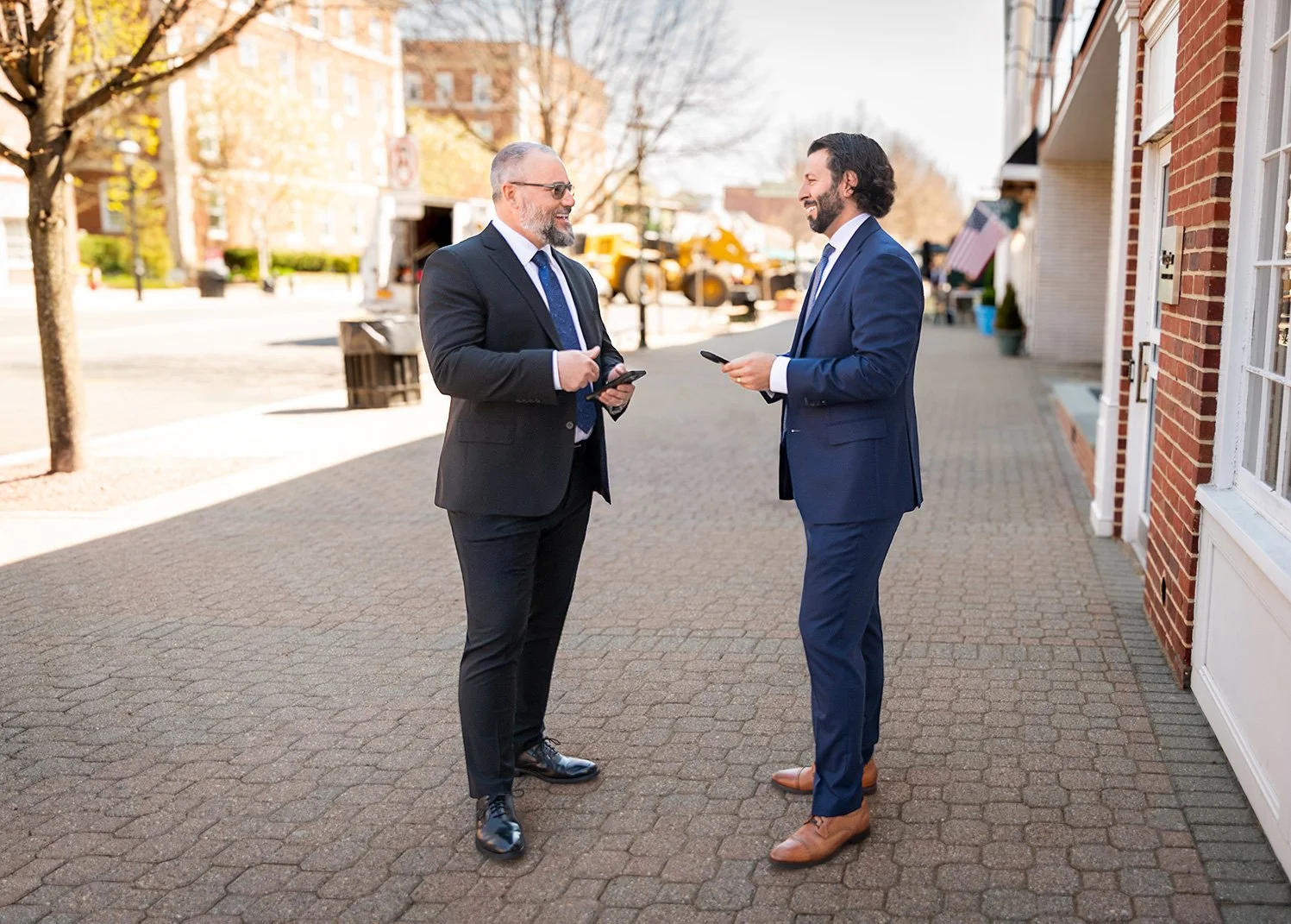 Two professional men in suits standing outdoors on a city sidewalk, smiling and talking while looking at their phones.