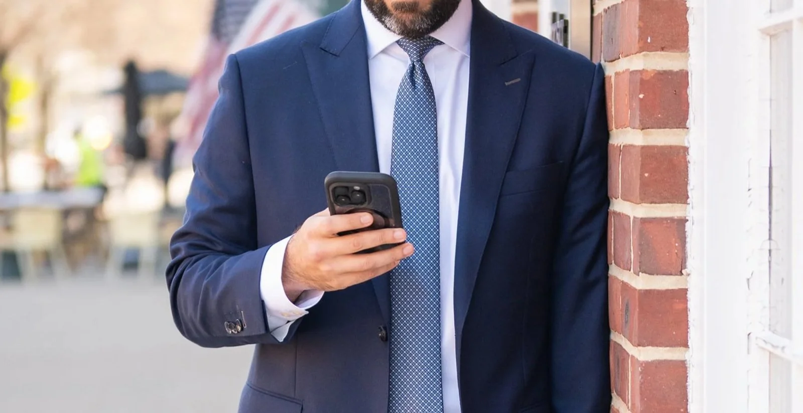 A man in a dark blue suit and tie is standing outside, leaning against a brick wall while looking at his smartphone.