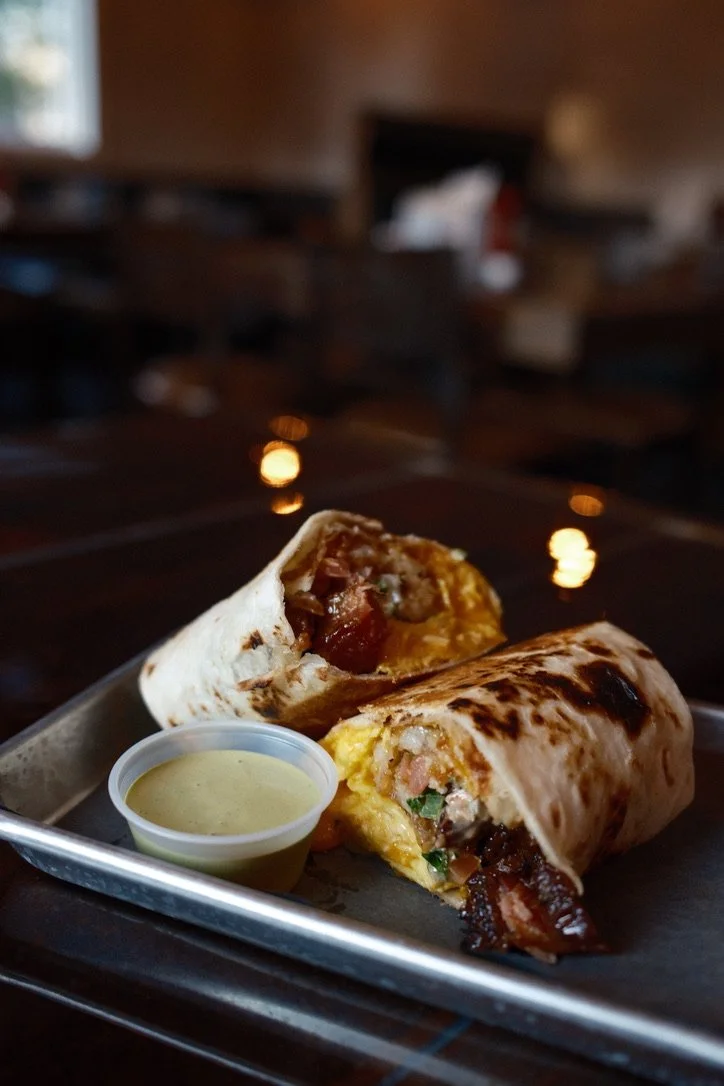 A burrito cut in half on a tray with a side of green sauce, inside with meat, cheese, and vegetables, served in a restaurant setting with warm lighting.