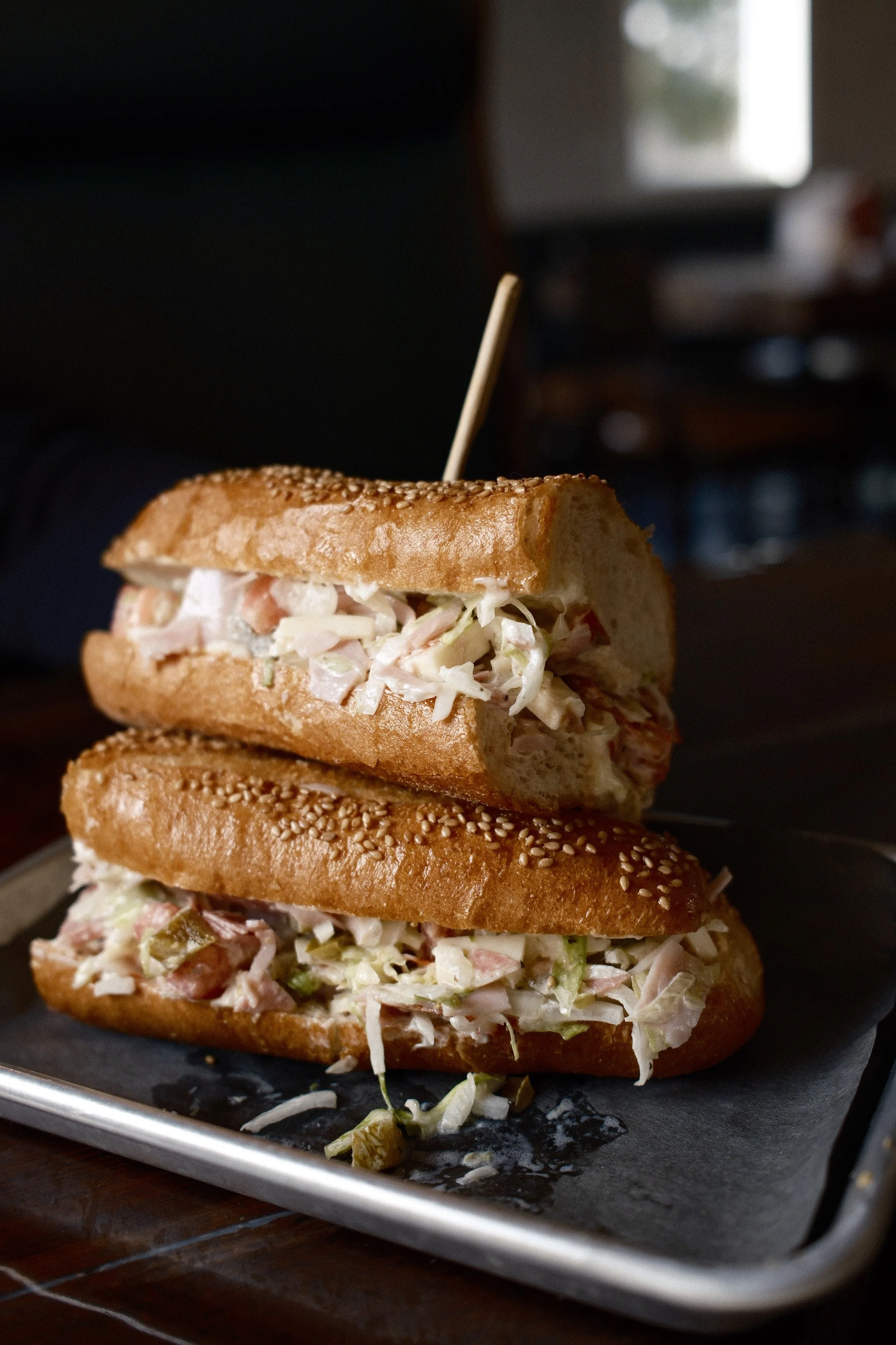 Two submarine sandwiches with sesame seed buns, filled with coleslaw and possibly ham or turkey, served on a black tray.