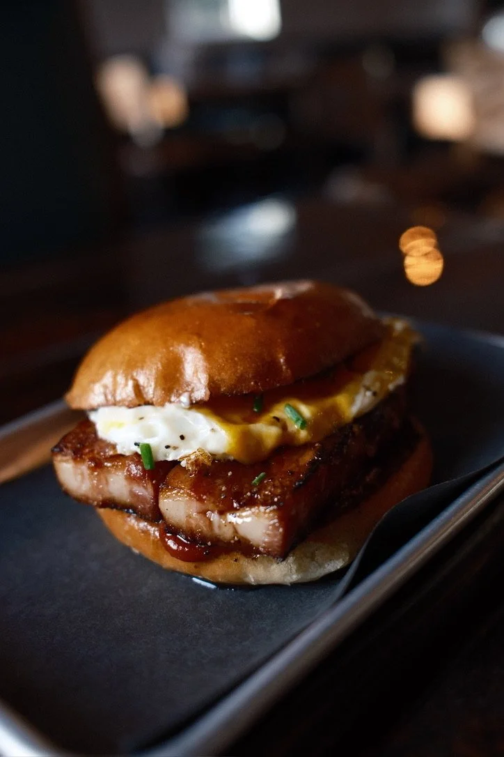 Close-up of a gourmet burger with a brioche bun, a thick slice of grilled pork belly, sunny-side-up egg with runny yolk, chives, and barbecue sauce, on a black plate.