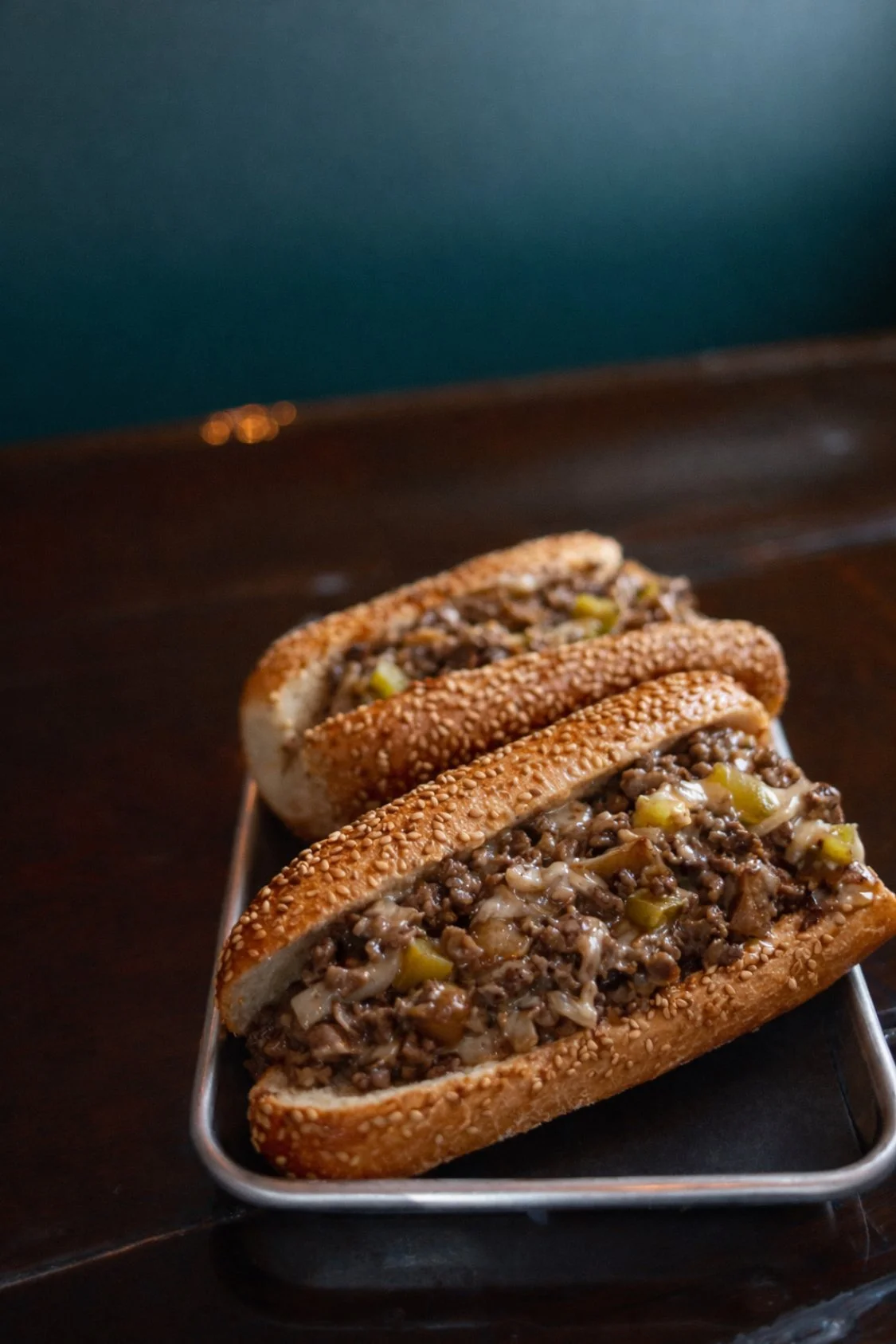 Three beef and onion sandwiches in sesame seed hoagie buns on a metal tray.