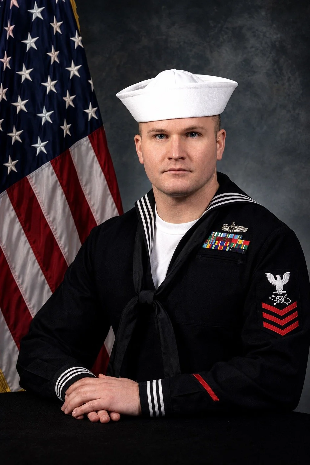 A man in a U.S. Navy sailor uniform with a white hat, sitting in front of an American flag, with a dark background.