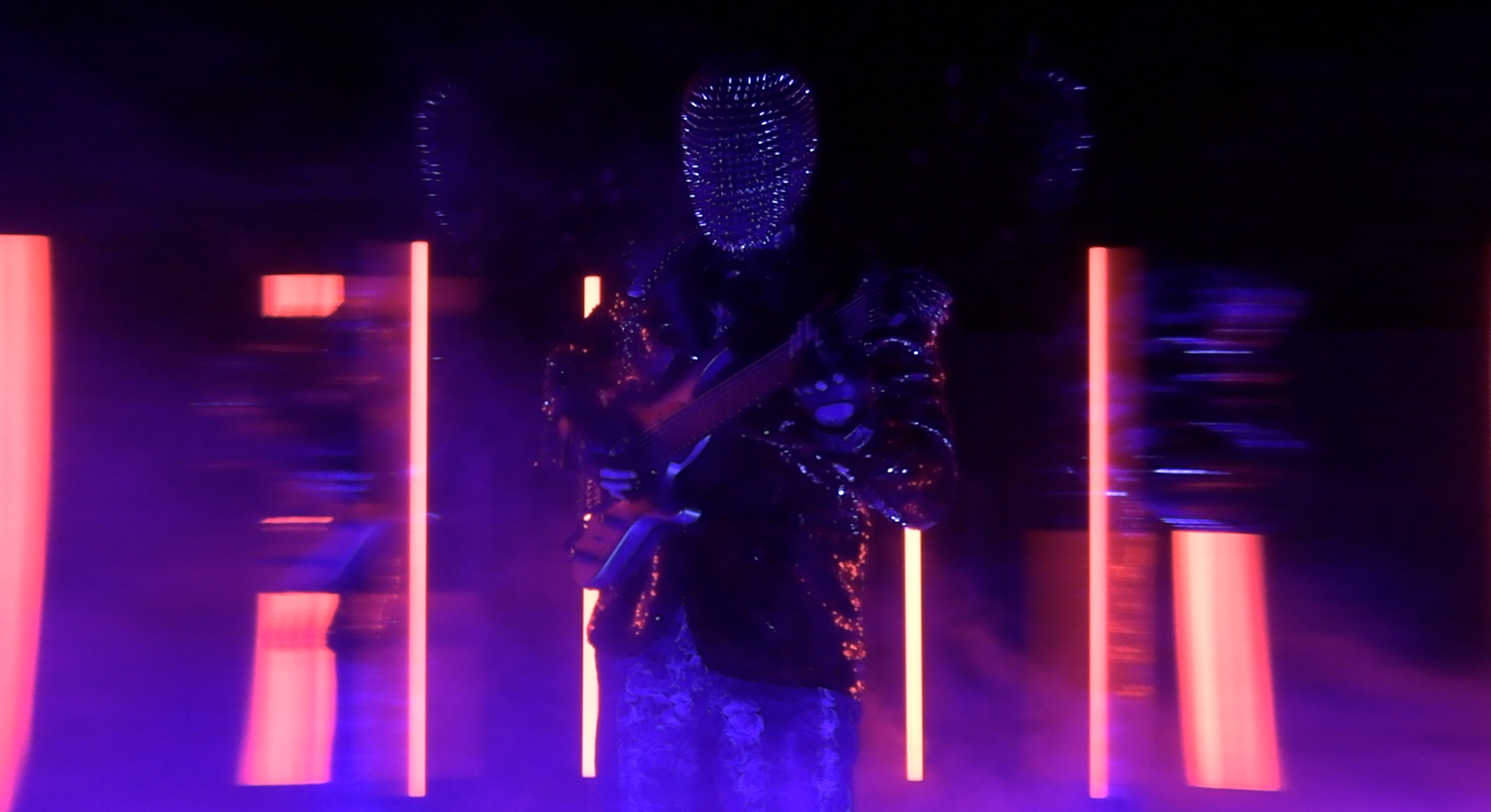 A person wearing a glowing, LED-lit mask playing an electric guitar in a dark room illuminated with purple and pink neon lights.