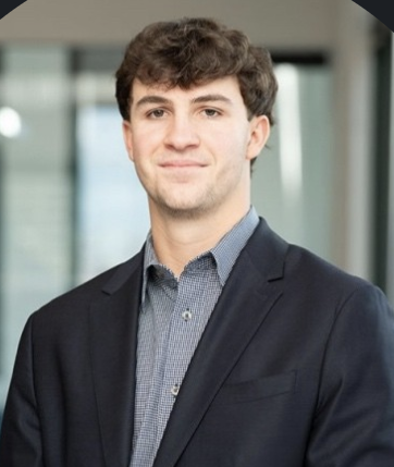 Young man with curly brown hair wearing a dark blazer and light blue shirt standing indoors with blurred windows in the background.