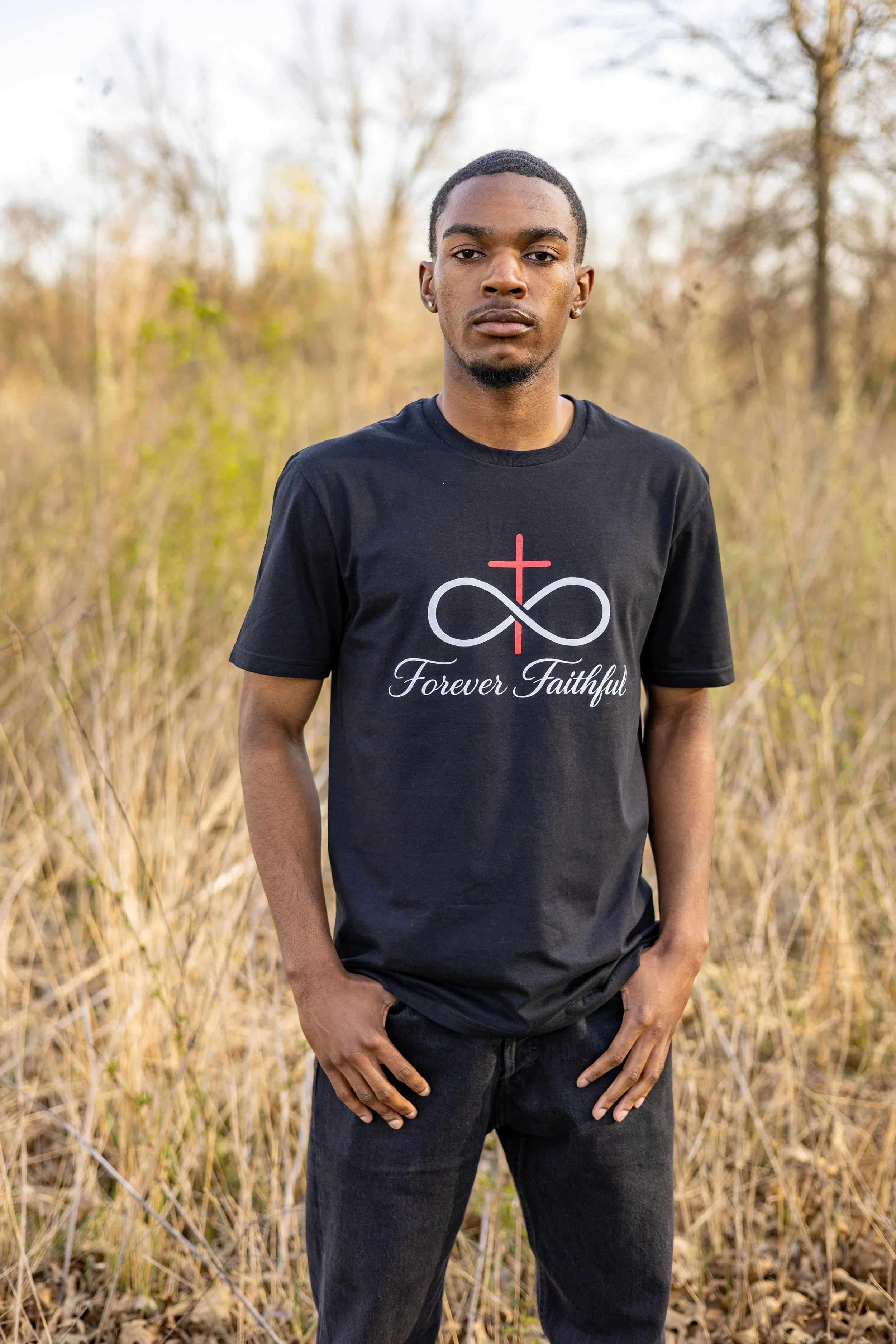 A young man standing outdoors in a field of tall grass and trees, wearing a black T-shirt with a faith-based logo and the words 'Forever Faithful' printed on it.