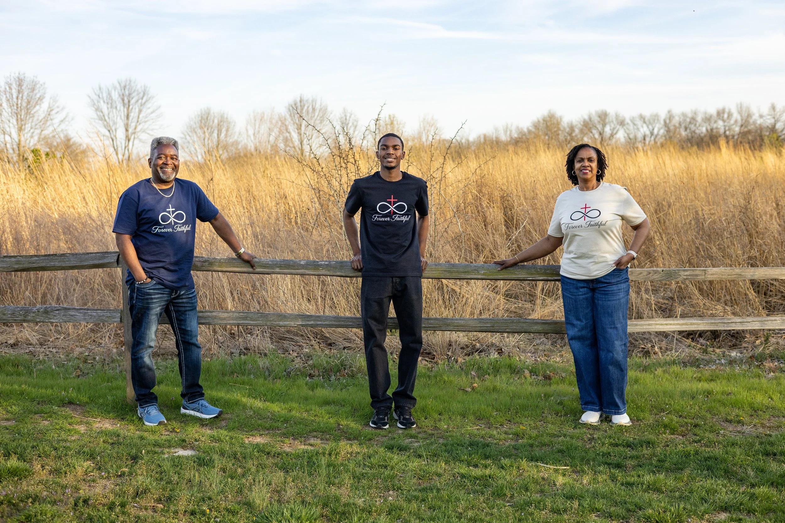 Three people standing outdoors near a wooden fence, smiling, with dry grass and trees in the background, all wearing shirts with religious symbols and text.