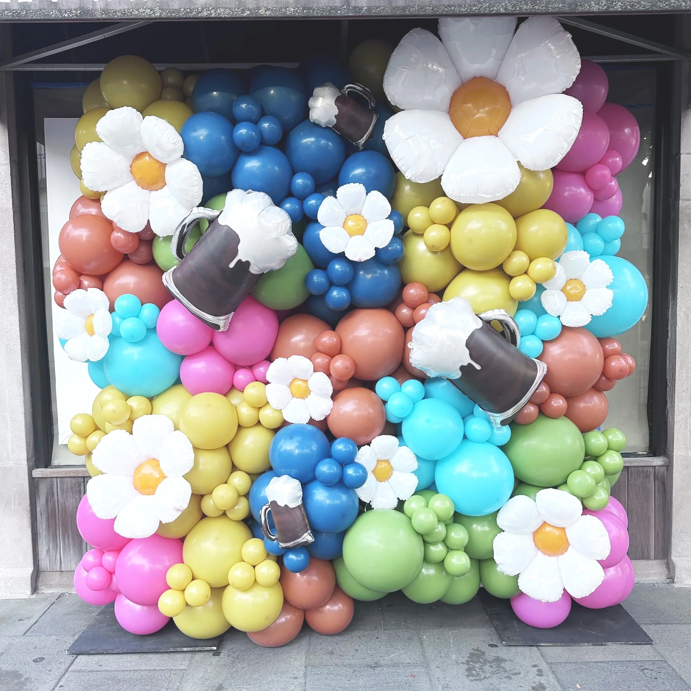 Colorful balloon display with large flower-shaped balloons, clusters of smaller balloons, and four large coffee mug-shaped balloons with whipped cream details, set against a window background.