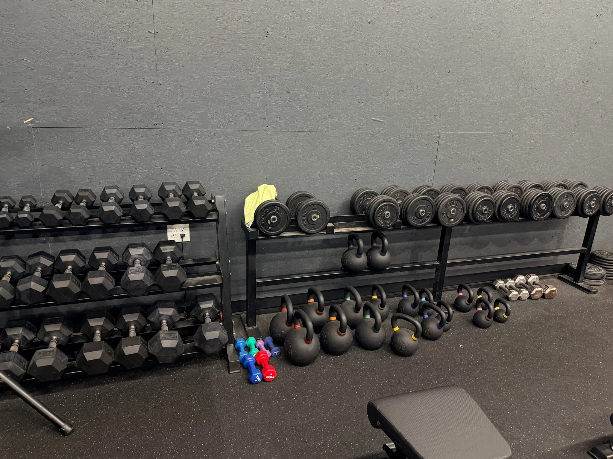 Gym equipment including black dumbbells on a rack, kettlebells on the floor, and smaller dumbbells on the ground, with a black workout bench in the foreground and a gray wall in the background.